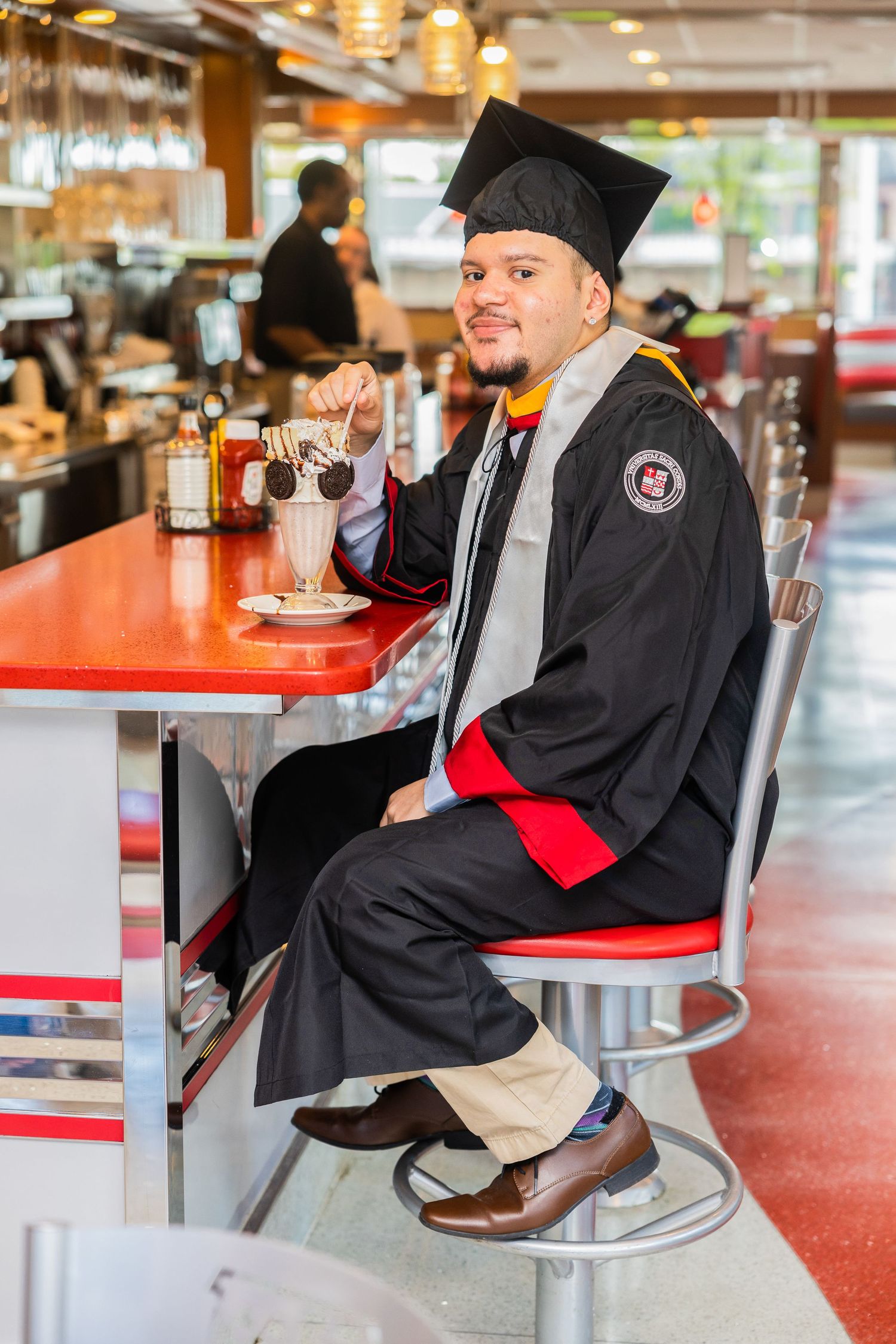 Graduate wearing graduation cap and gown sits at a retro diner counter with red stools.
