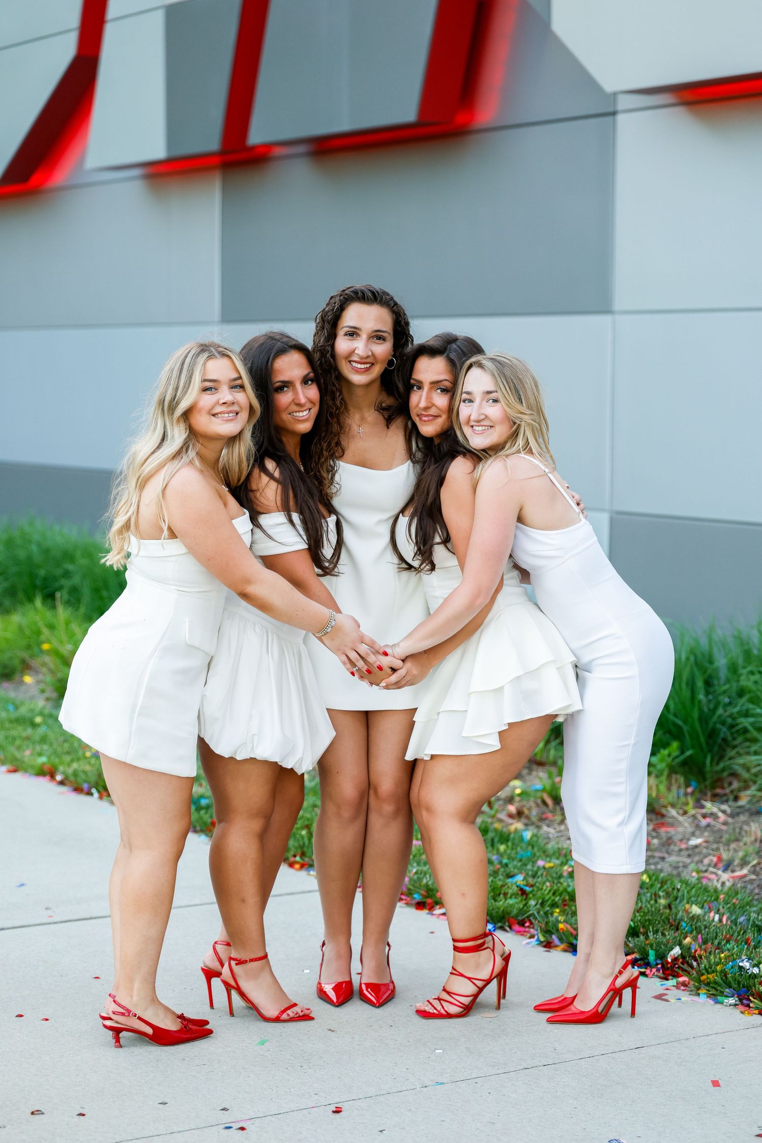 Group in white dresses shows off their matching red high heels while posing together outdoors.