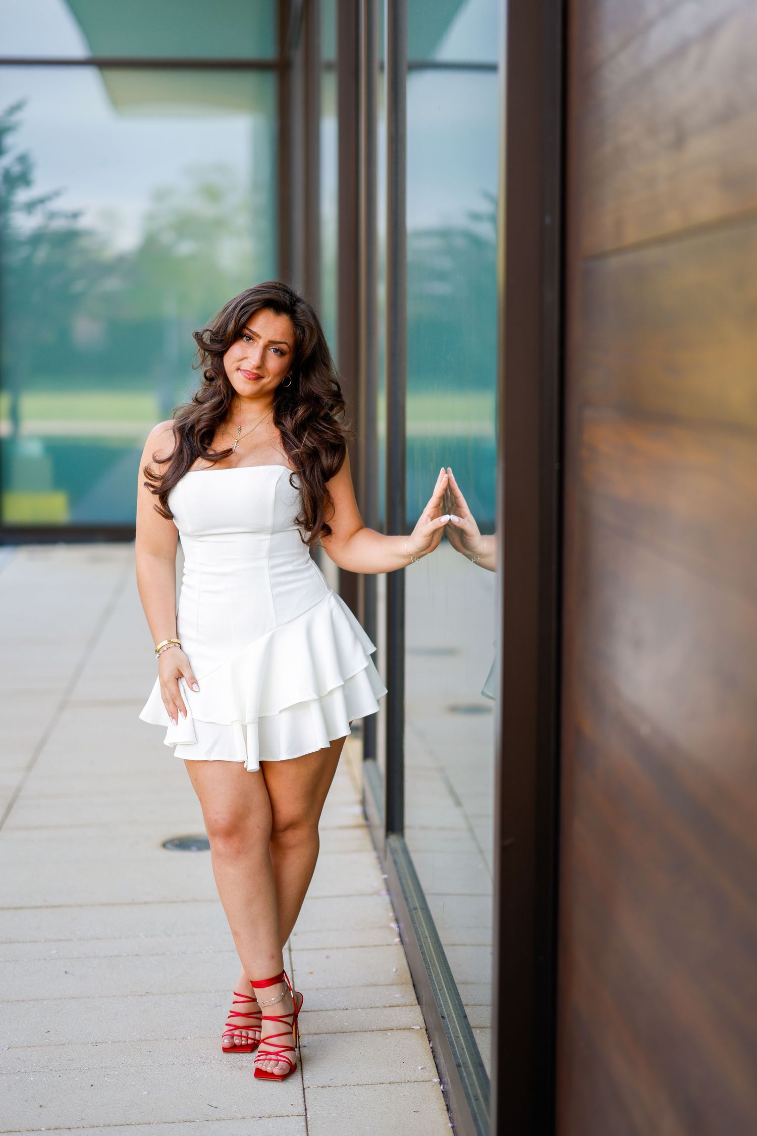 Person in white dress poses stylishly with red strappy heels near glass building wall.