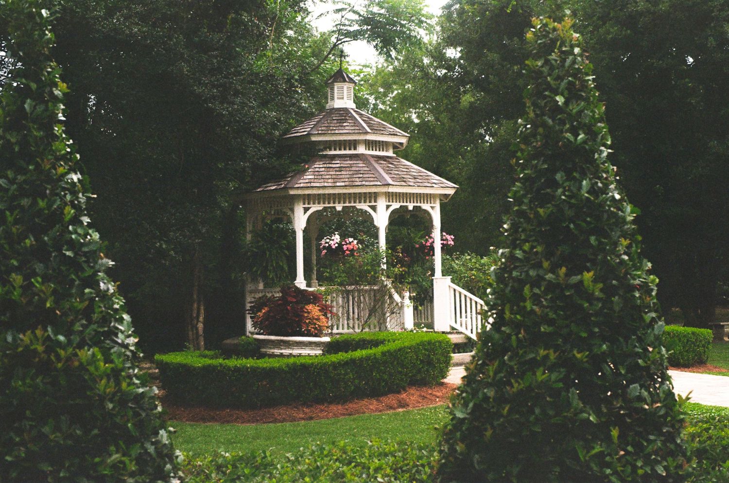 A white wooden gazebo surrounded by manicured bushes and trees in a lush garden setting.