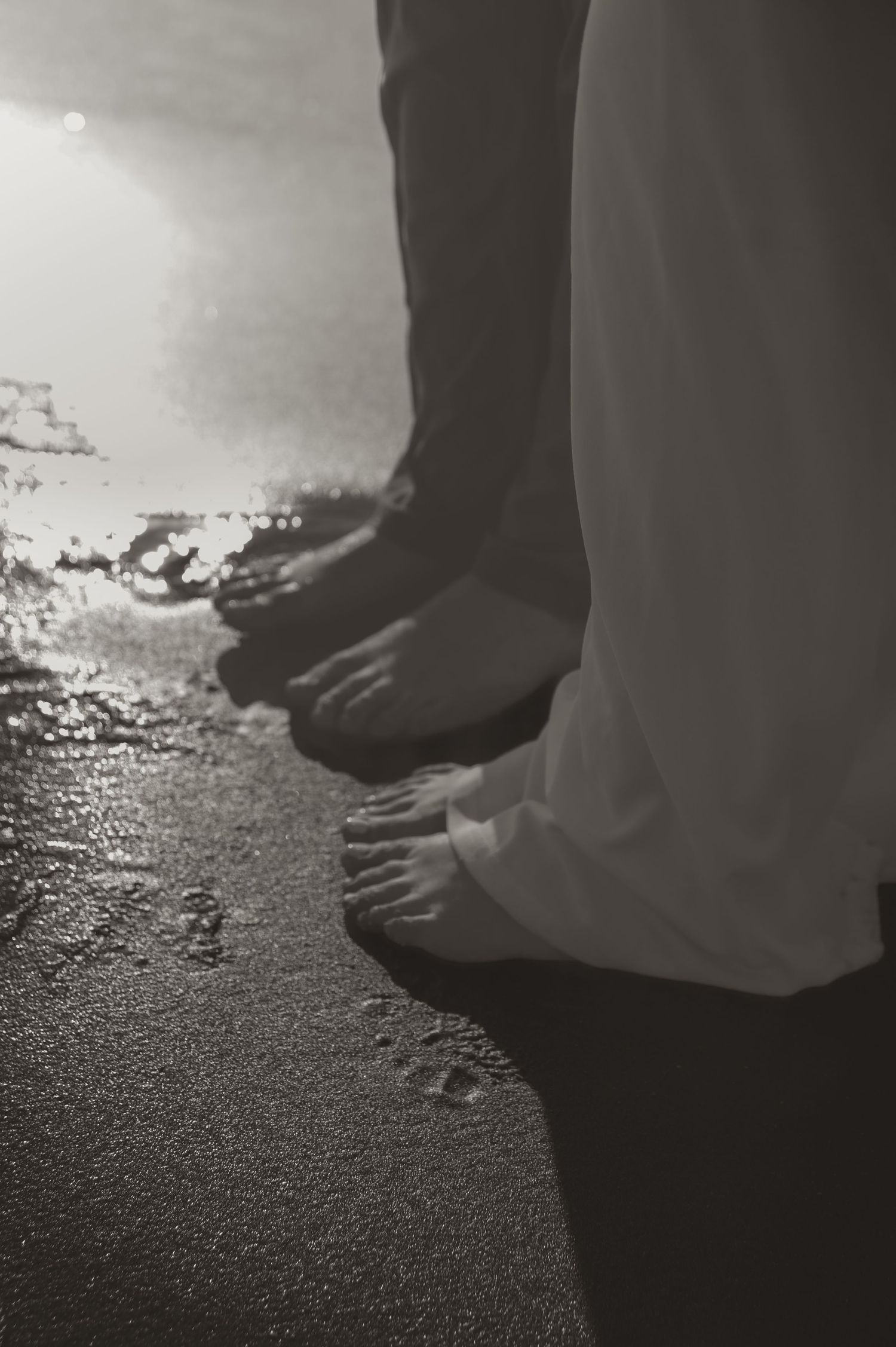 A black and white photo shows barefoot people standing at the edge of wet sand on a beach.