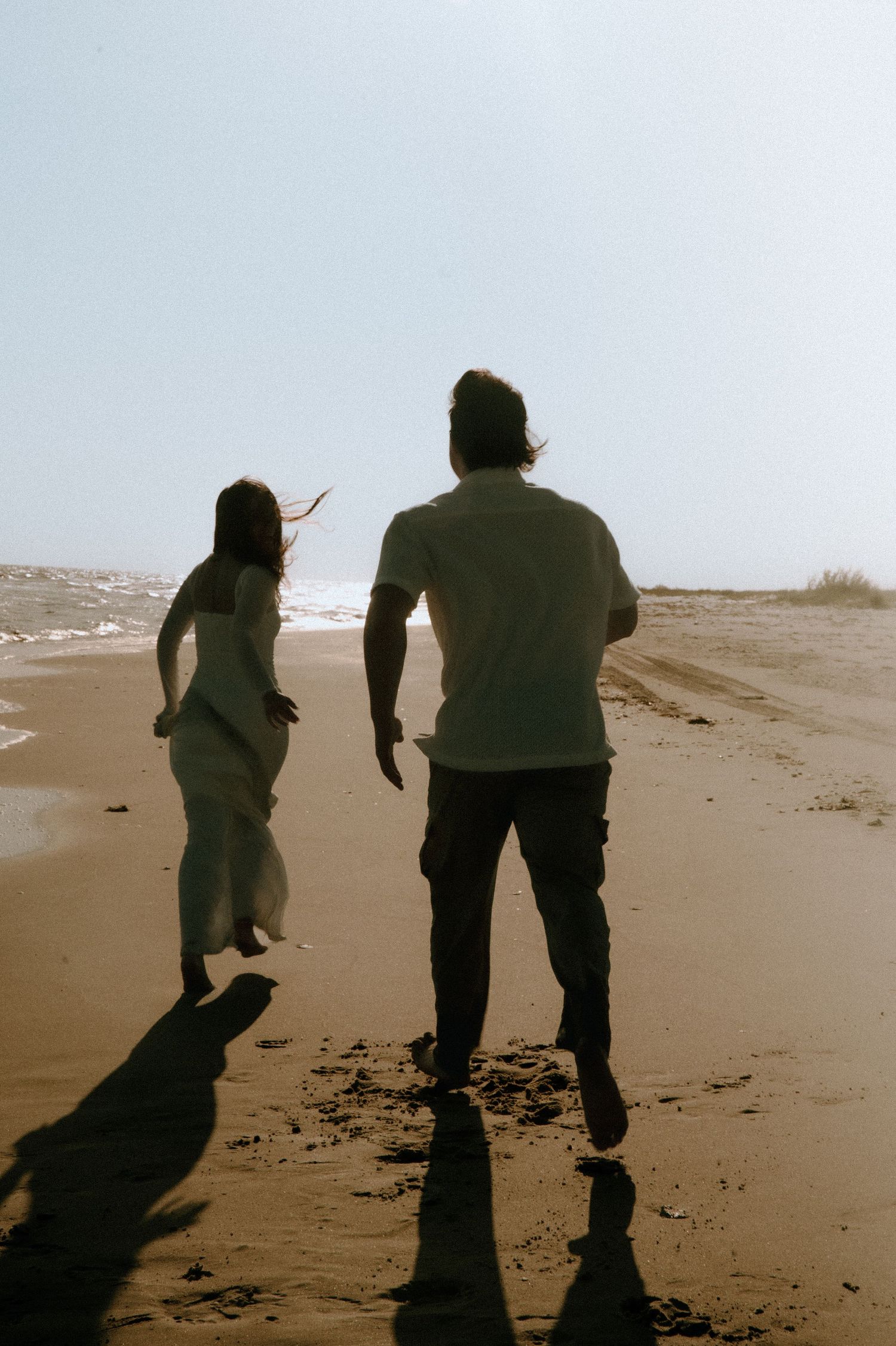 Two silhouetted figures walking along a beach shoreline at dusk casting long shadows on the sand.