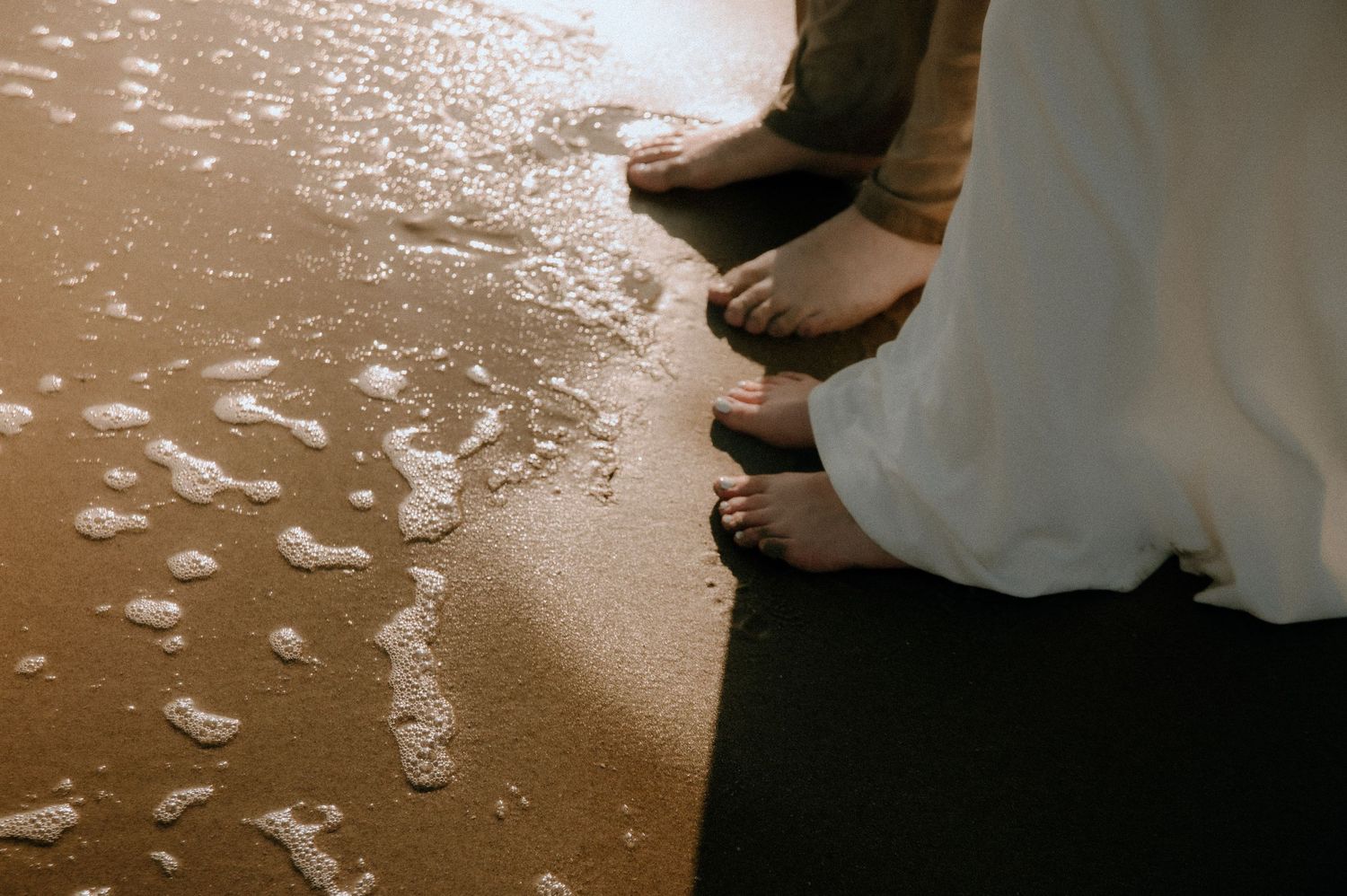 Bare feet of the couple stand on a sandy beach with the ocean water creating a gentle shoreline nearby.