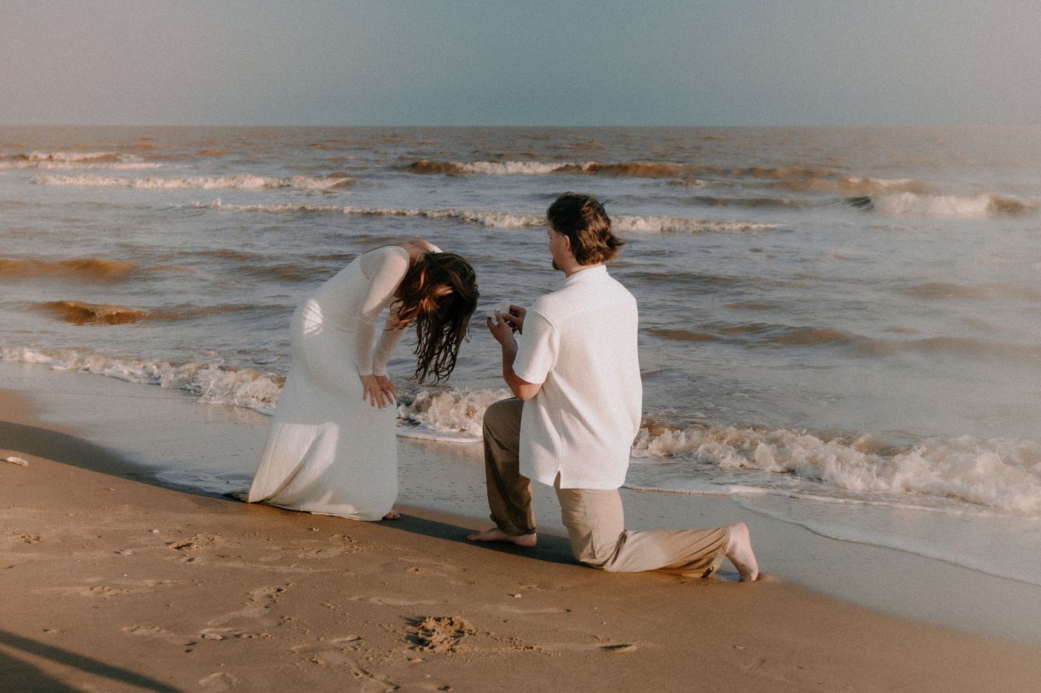 A couple wearing white sits near the ocean waves at the beach during an intimate beachside moment.