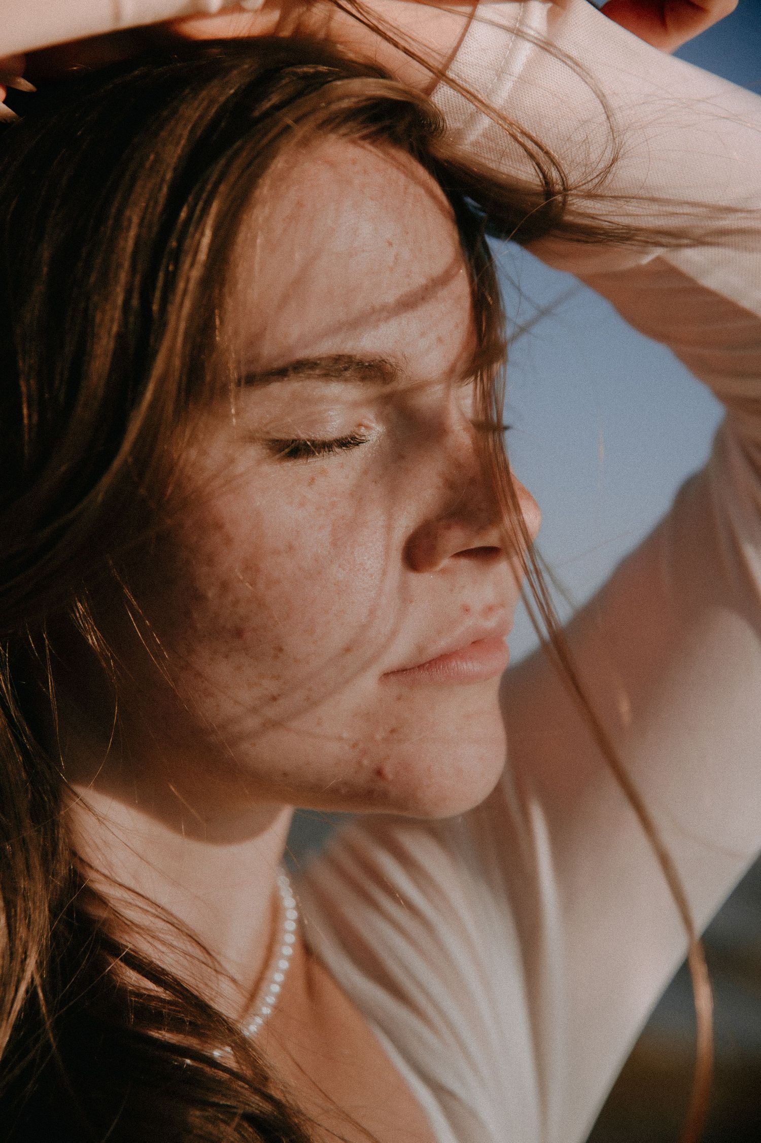 Close up portrait of person with eyes closed in sunlight, head tilted back with windswept brown hair.