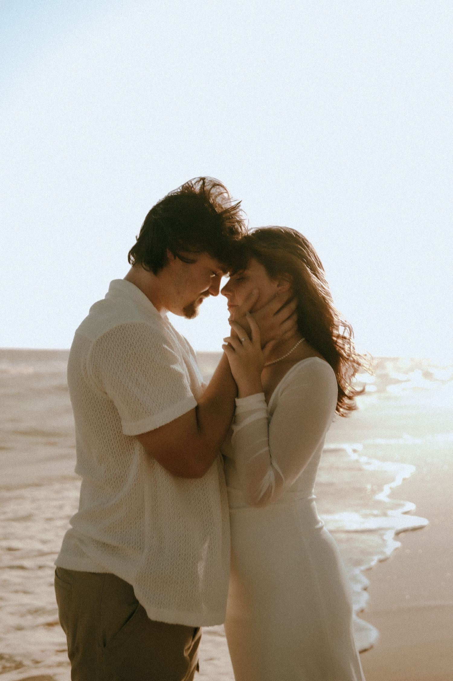 A romantic couple shares an intimate moment at sunset on a beach, dressed in white outfits as waves crash behind them.