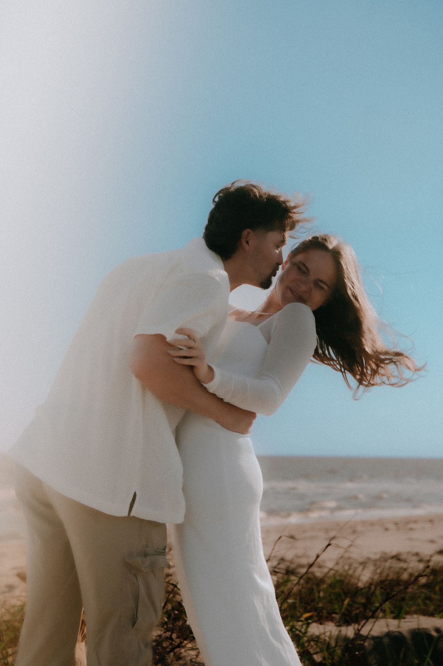 Romantic couple in white outfits embrace on a sunny beach with wind-swept hair and soft lighting.