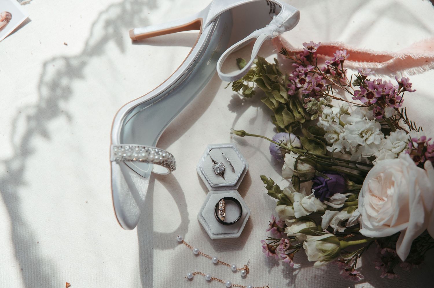 Wedding details flatlay with white heels, engagement rings, and delicate pink roses in soft natural lighting.