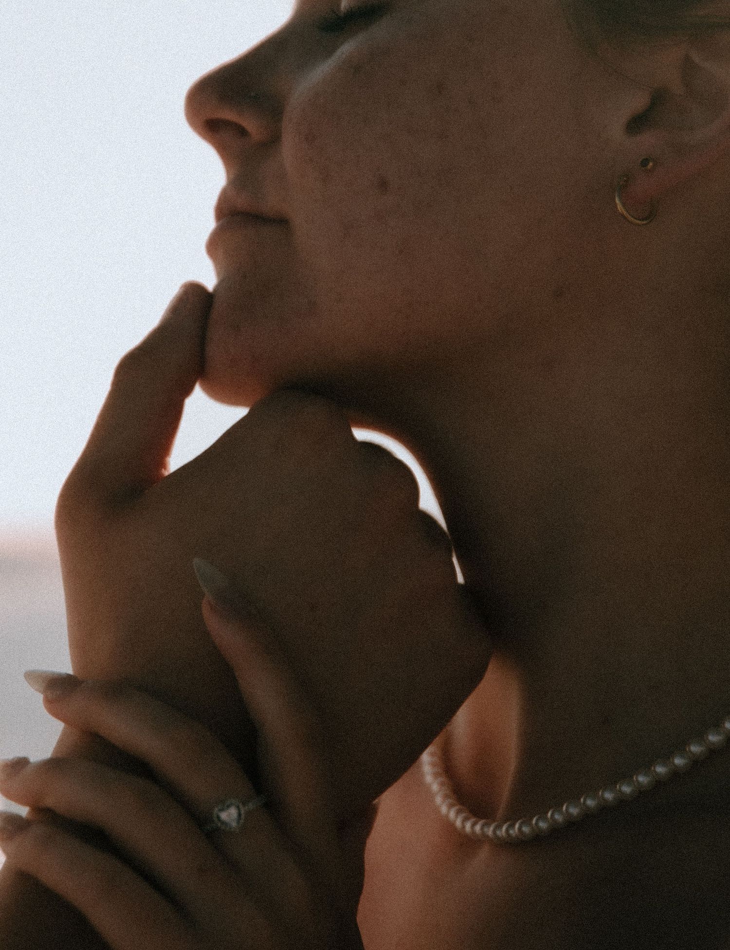 Close-up artistic portrait showing hands gently touching neck and chin, wearing delicate jewelry in soft lighting.