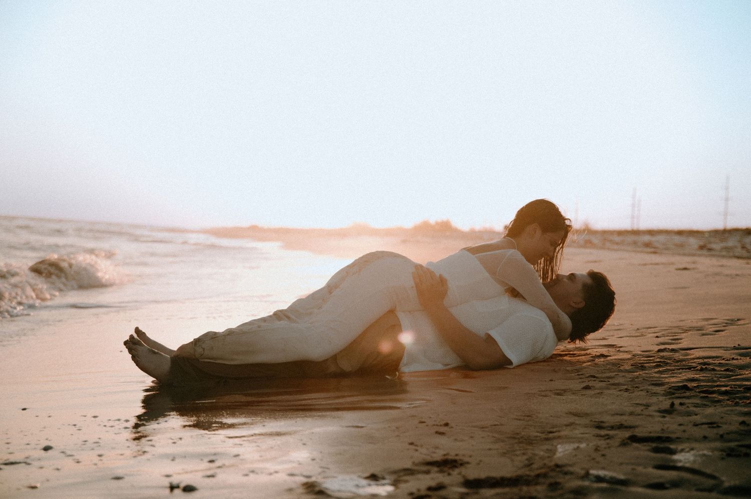 Two people in white clothing lying together on a sandy beach during golden hour sunset with dreamy lighting.
