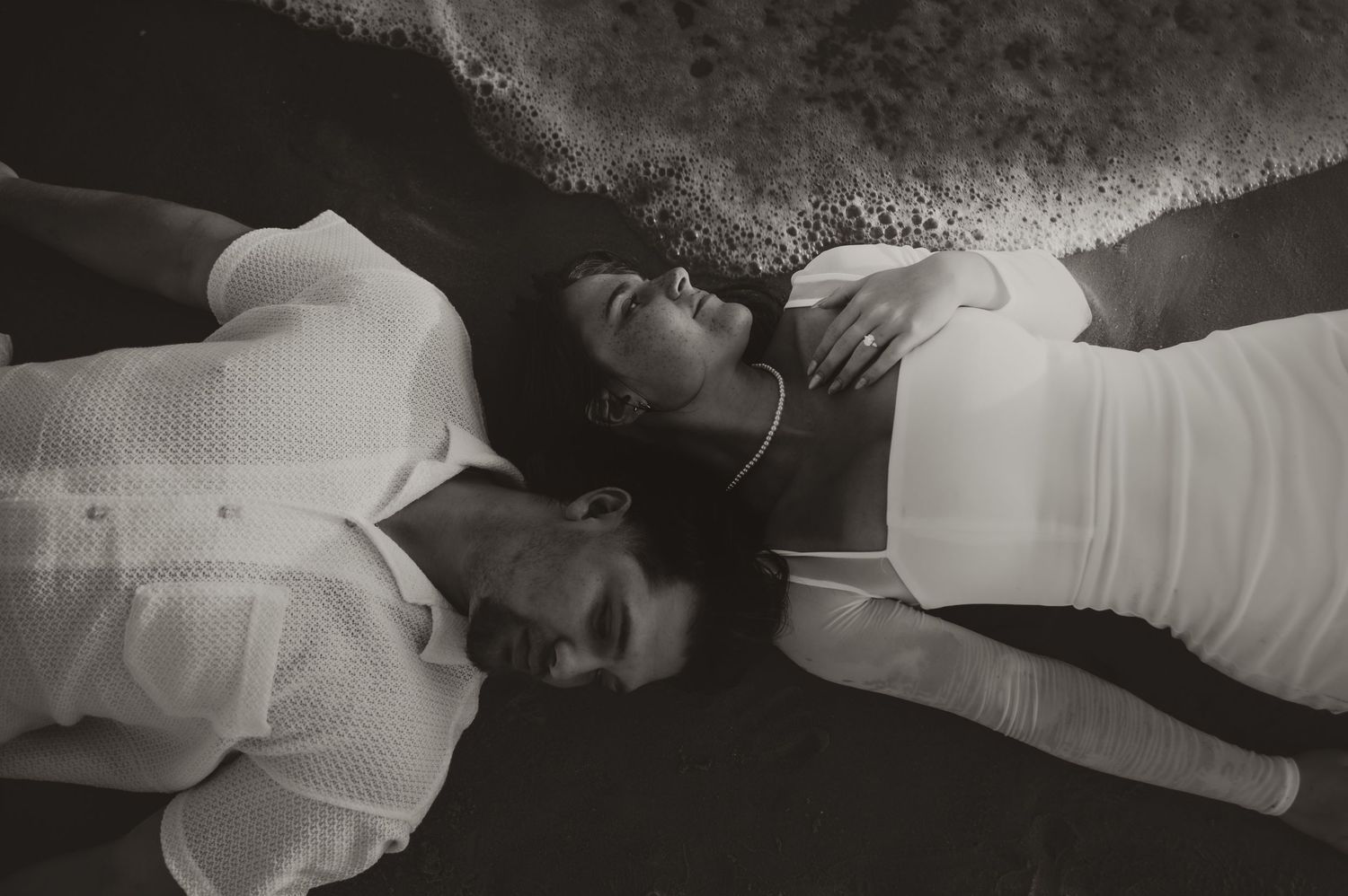 Two people in white clothing lay on a dark surface with their heads together in a black and white artistic portrait.