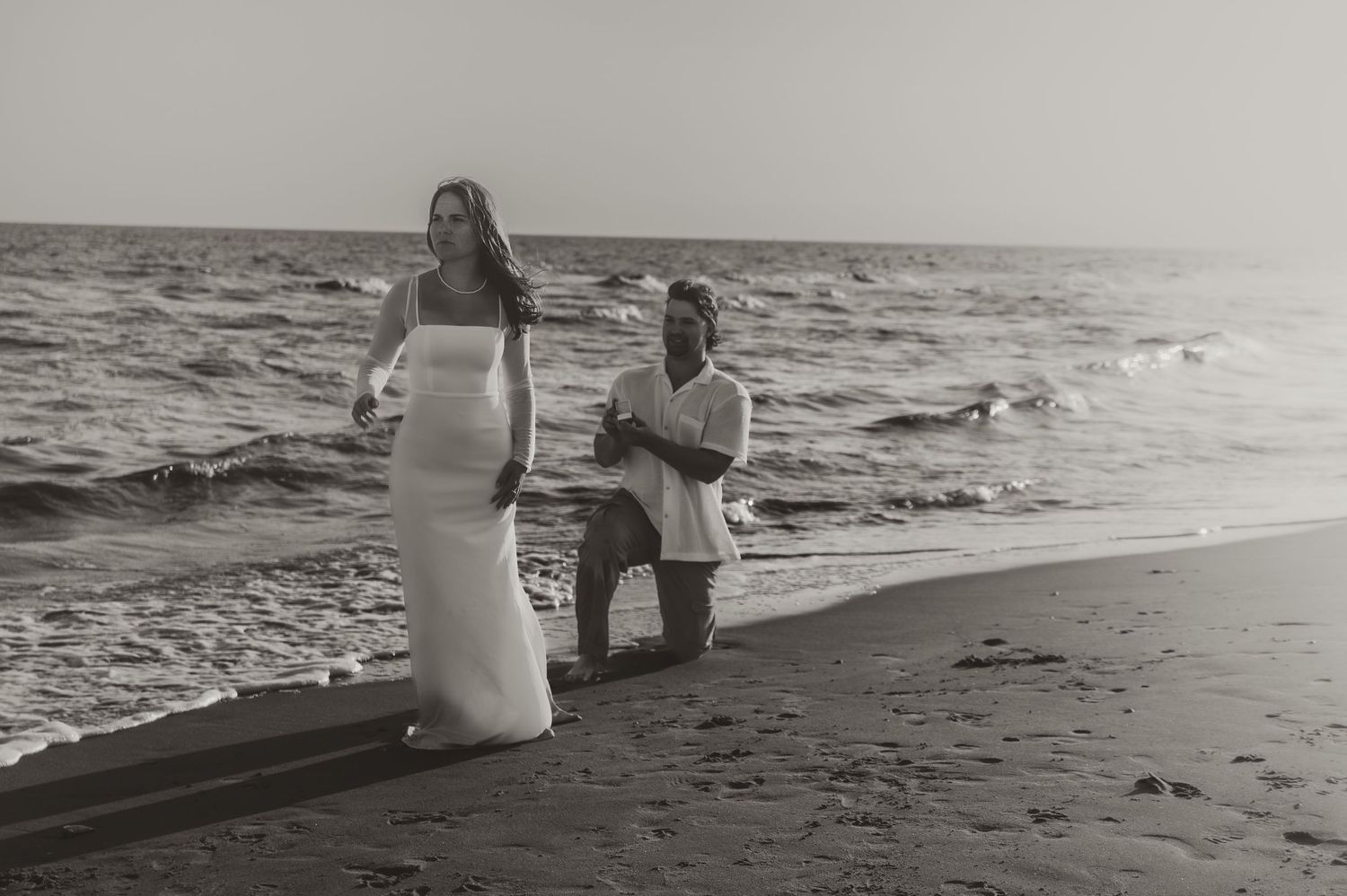 Two people in light colored clothing stand along the beach shoreline in a black and white photograph.