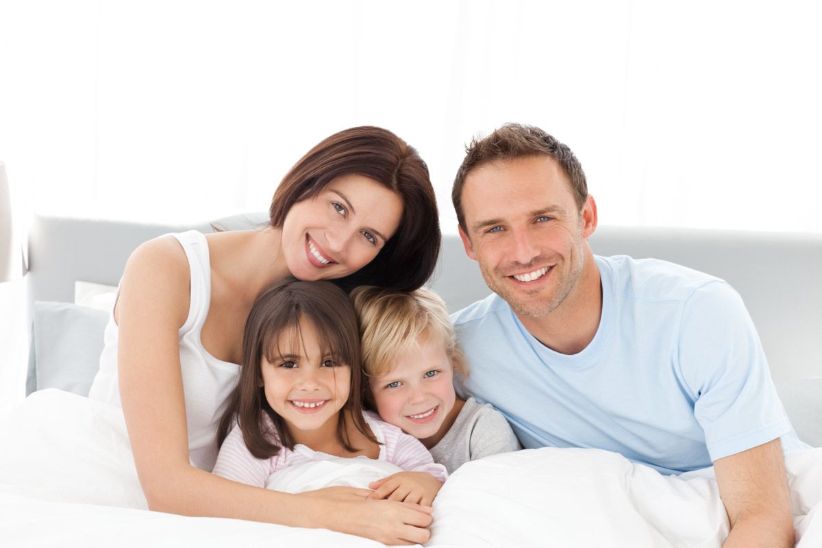 Smiling family of four in natural light sitting on bed.