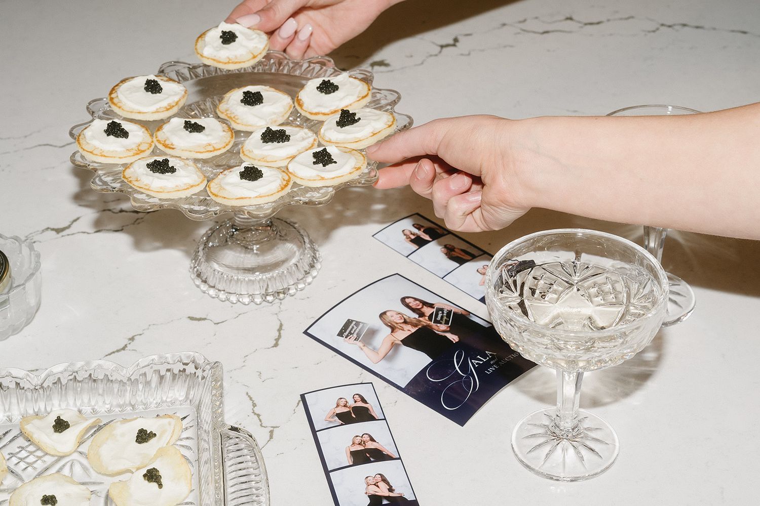 A hand places black and white decorated cookies onto a decorative glass cake stand and pedestal display.