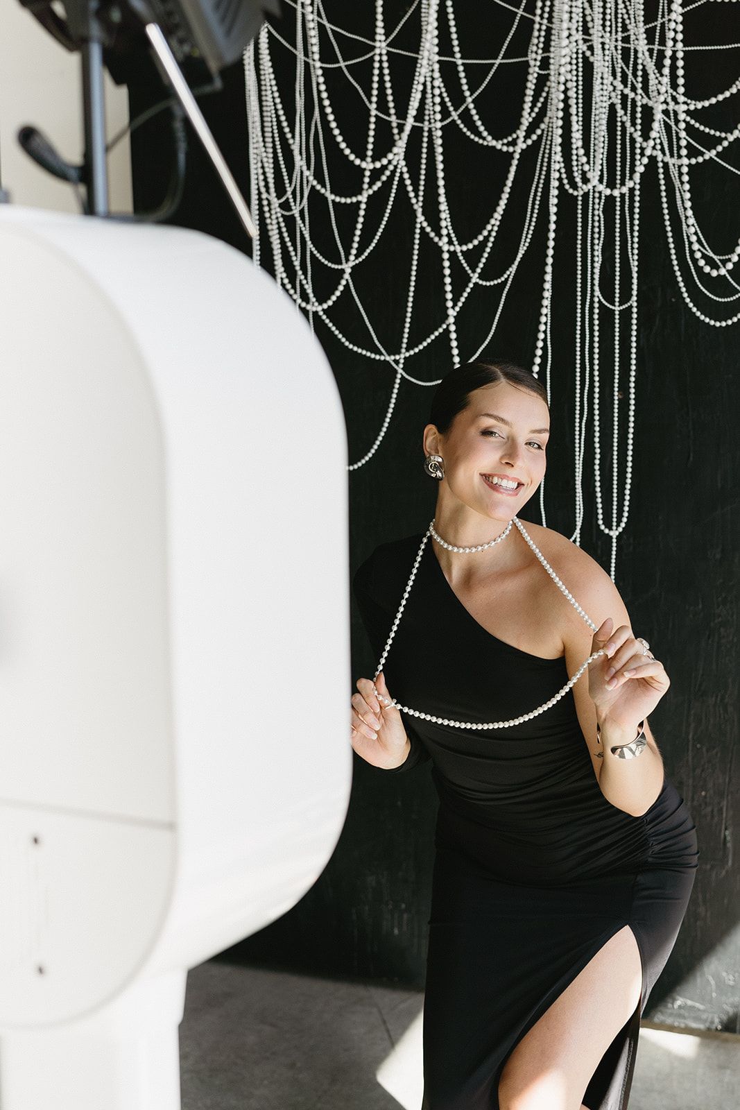 Black-clad person poses playfully against dark background with decorative hanging pearl chains and white curved element.