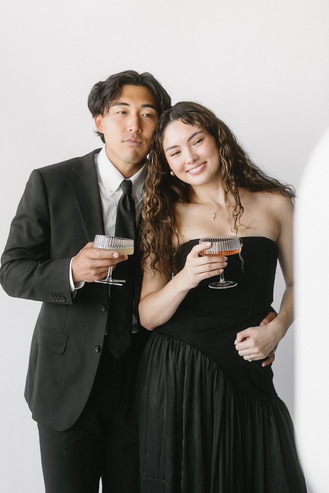 A couple in formal attire holding wine glasses poses together against a white backdrop in three similar shots.