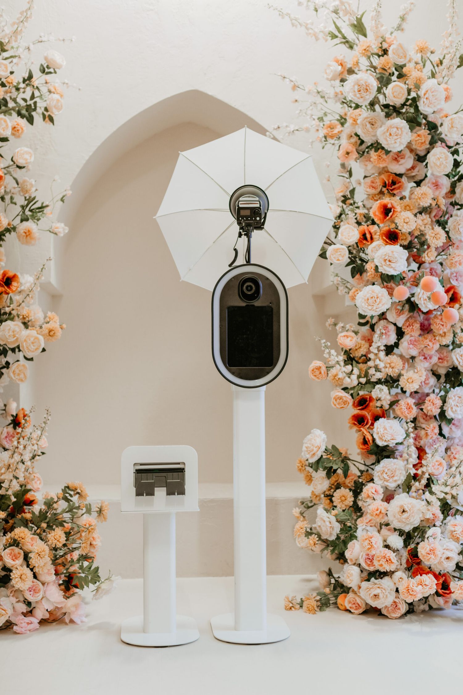 Photo booth setup with white pedestals, umbrellas, and elegant peach floral arrangements against an arched white backdrop.