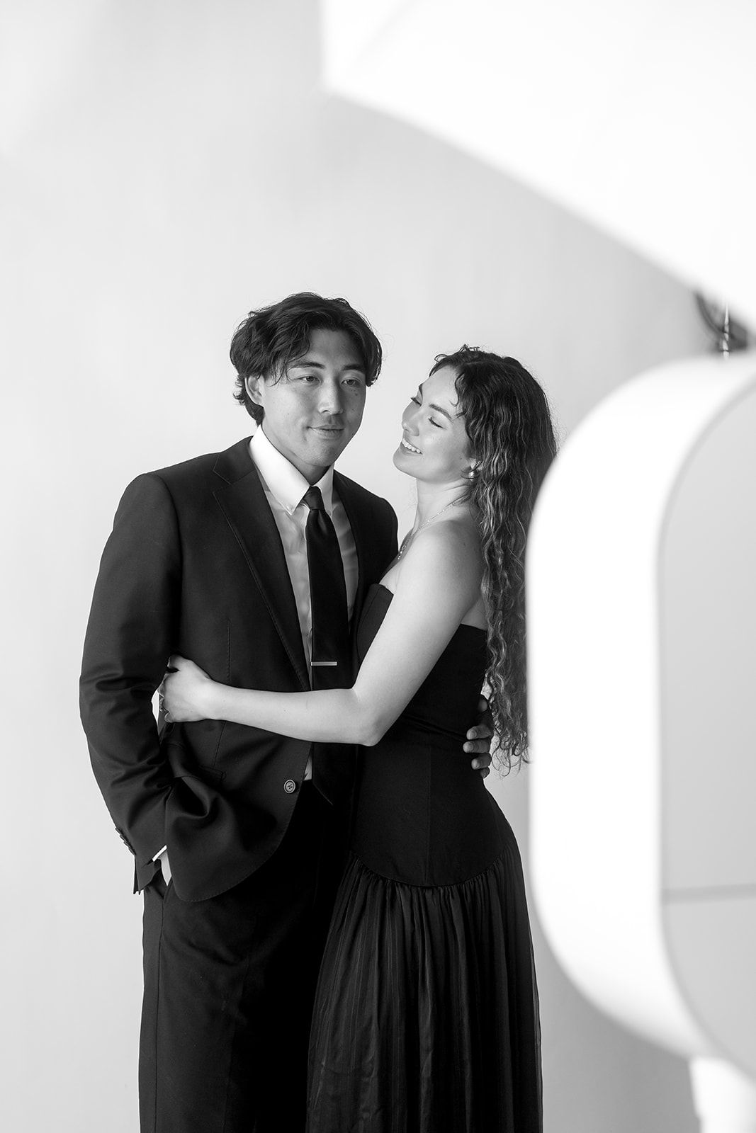 A romantic black and white portrait of a couple in formal attire embracing against a light background.