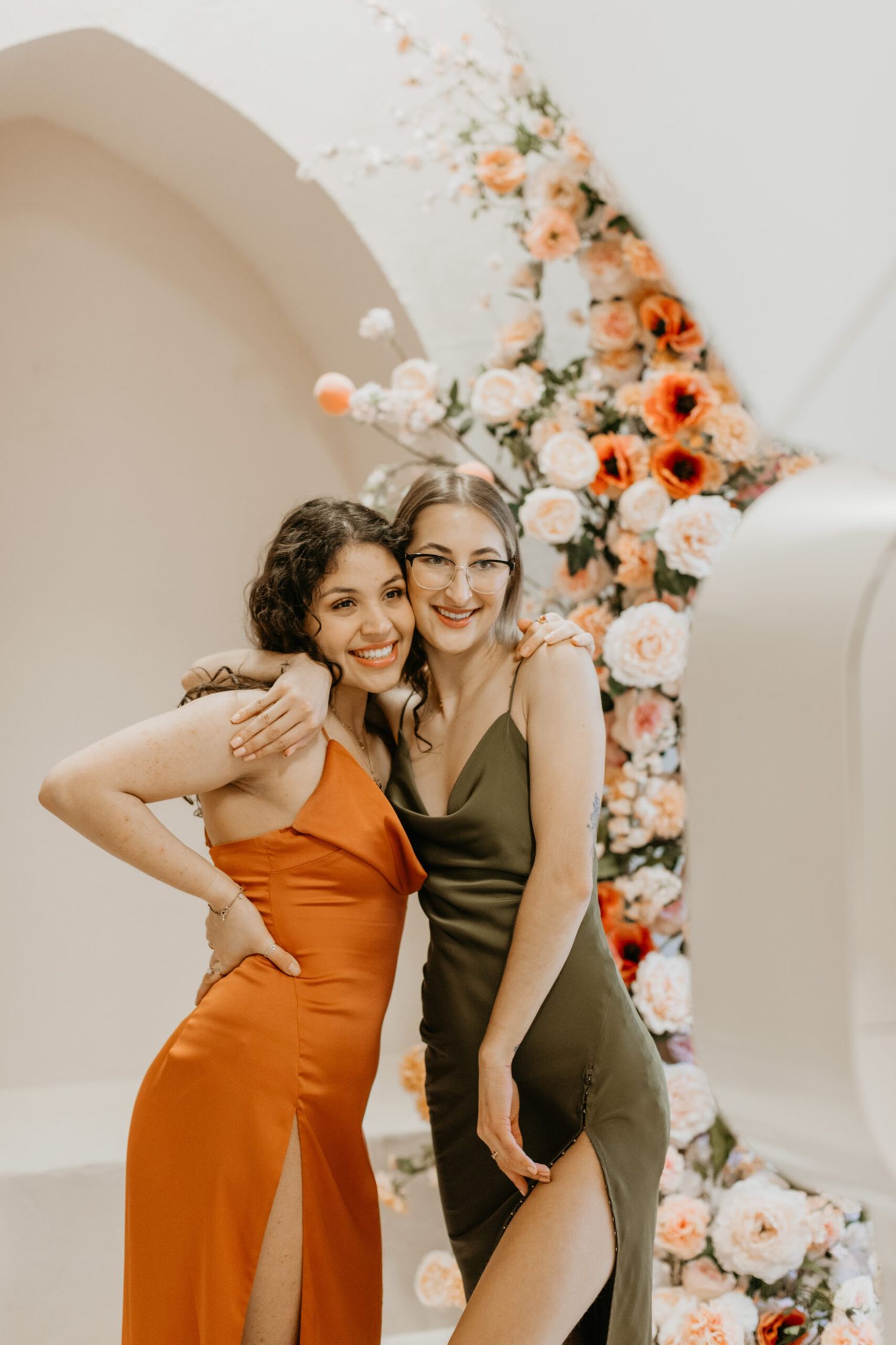 Two friends in contrasting dresses share candid moments against a white wall with peach flowers.