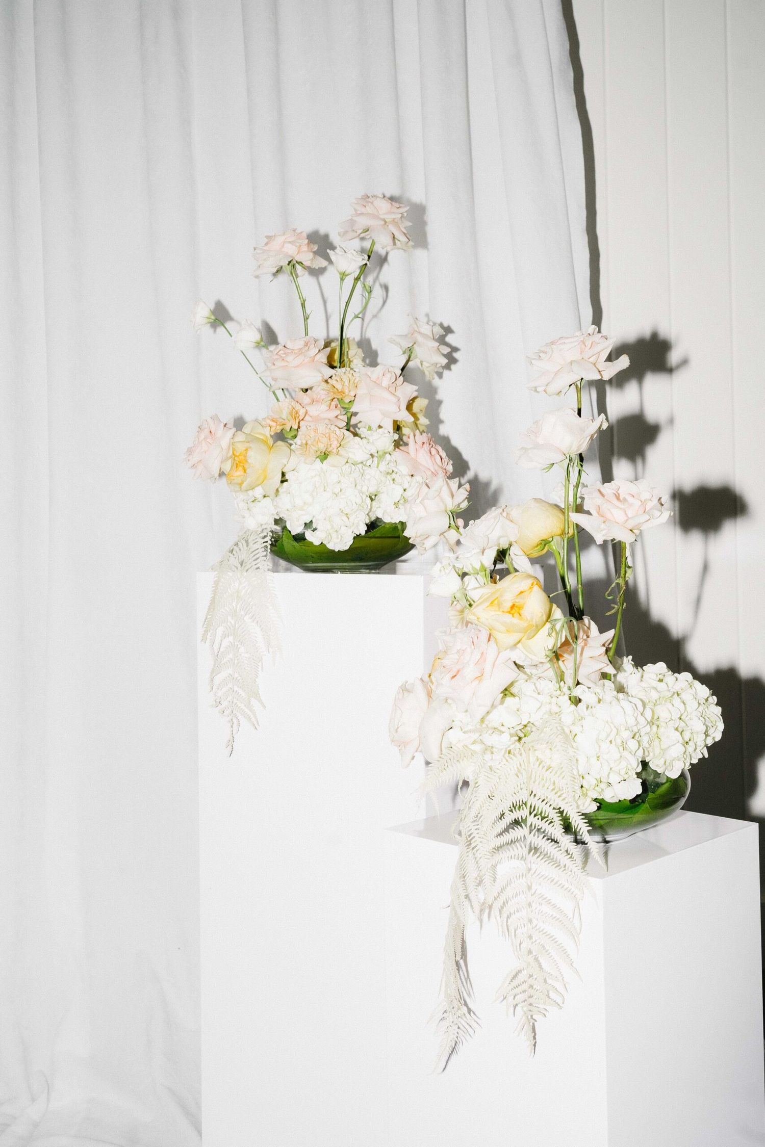 White ceramic vases filled with delicate pastel pink and cream floral arrangements against a white textural background.