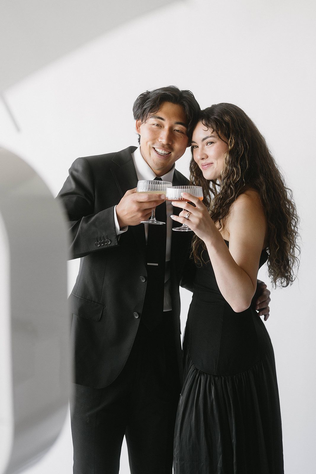 A couple in elegant black attire share a toast with drinks while smiling together against a white wall.