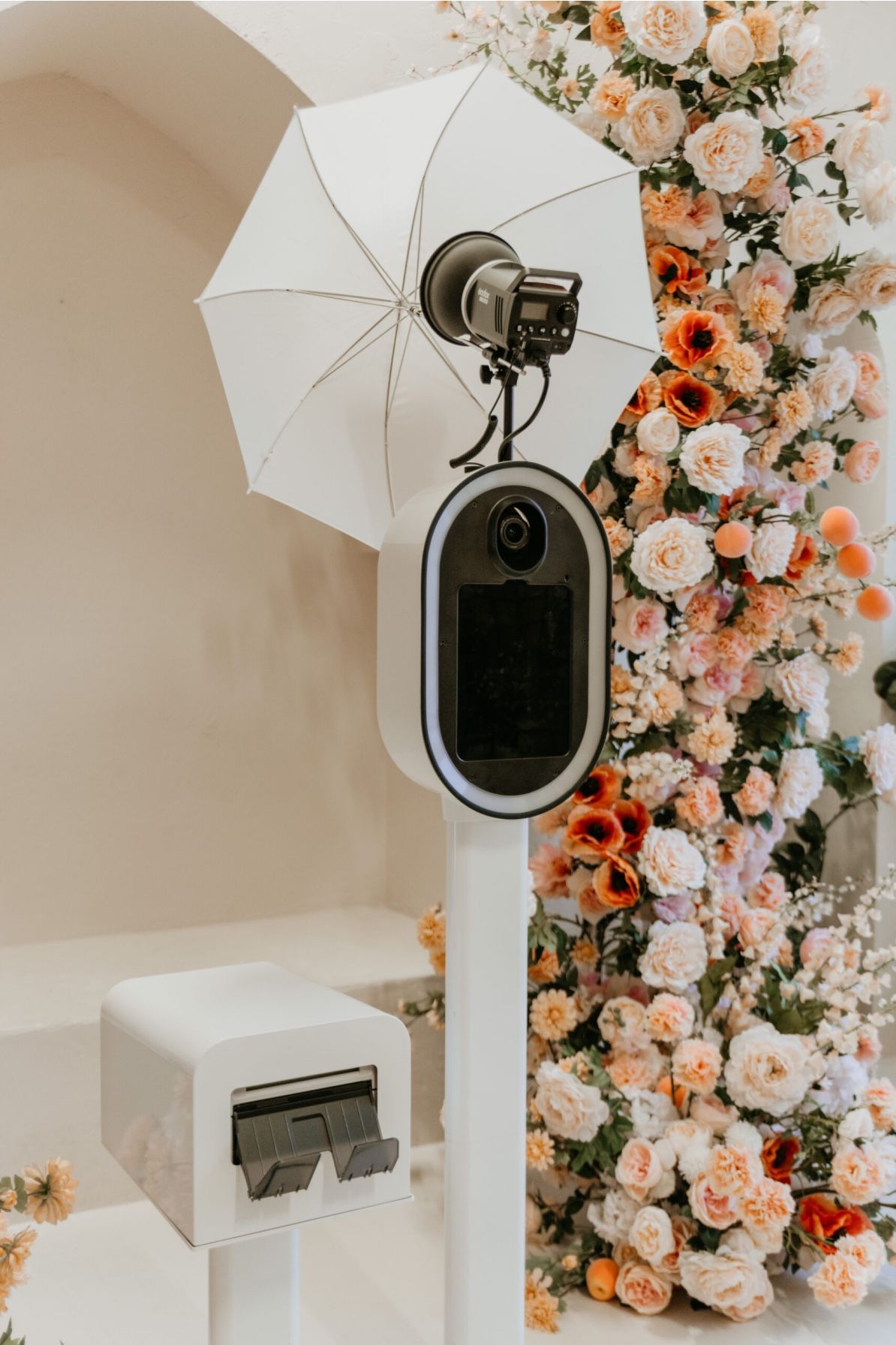 Studio photography setup with white umbrellas and camera against a wall decorated with peach and coral rose floral arrangements.