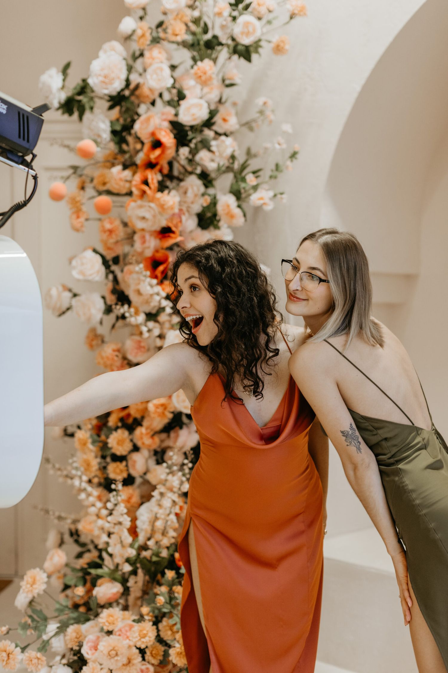 Candid photography sequence shows two friends laughing together in front of a floral arch.
