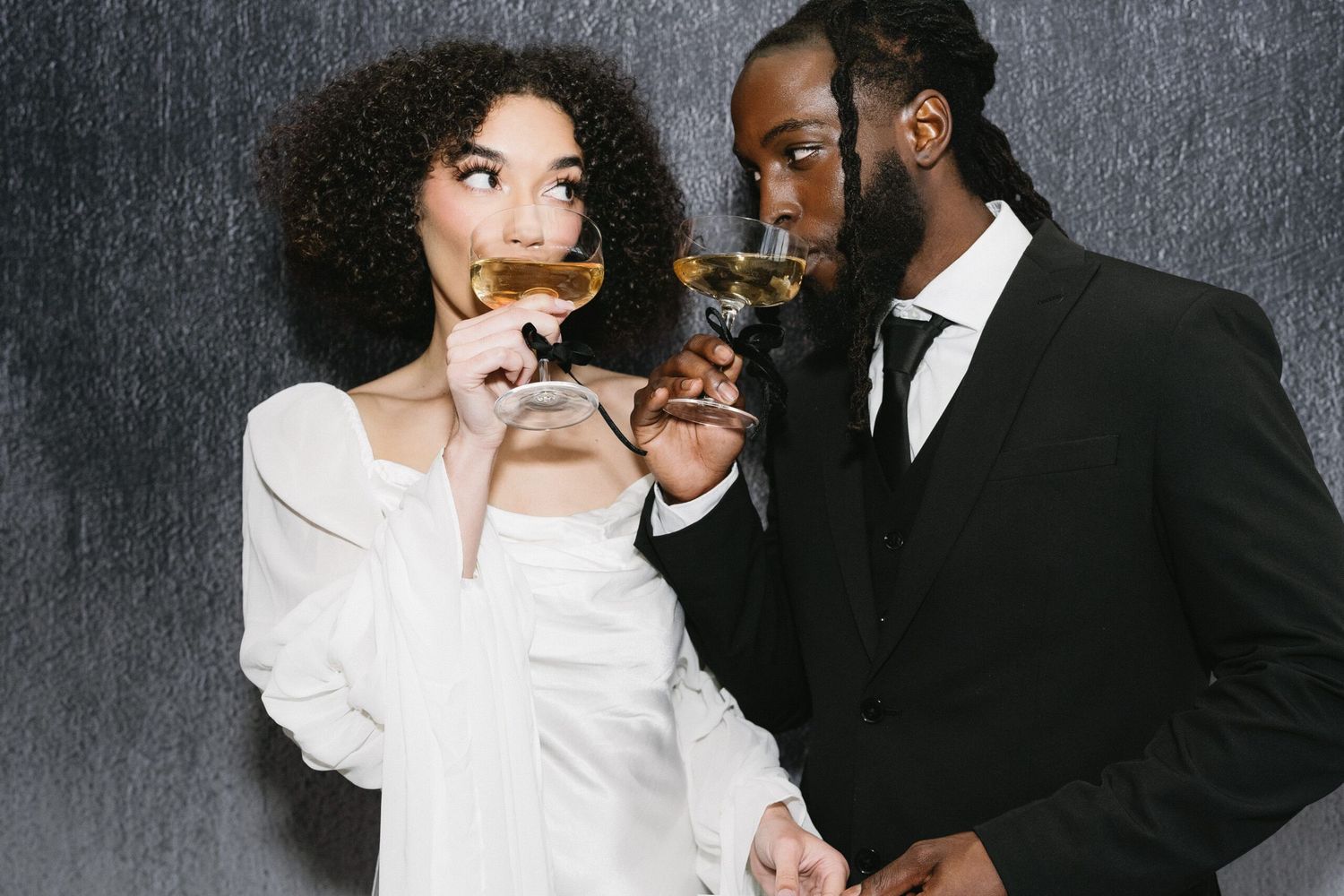 Two people in formal attire share a romantic moment with champagne against a dark textured background.