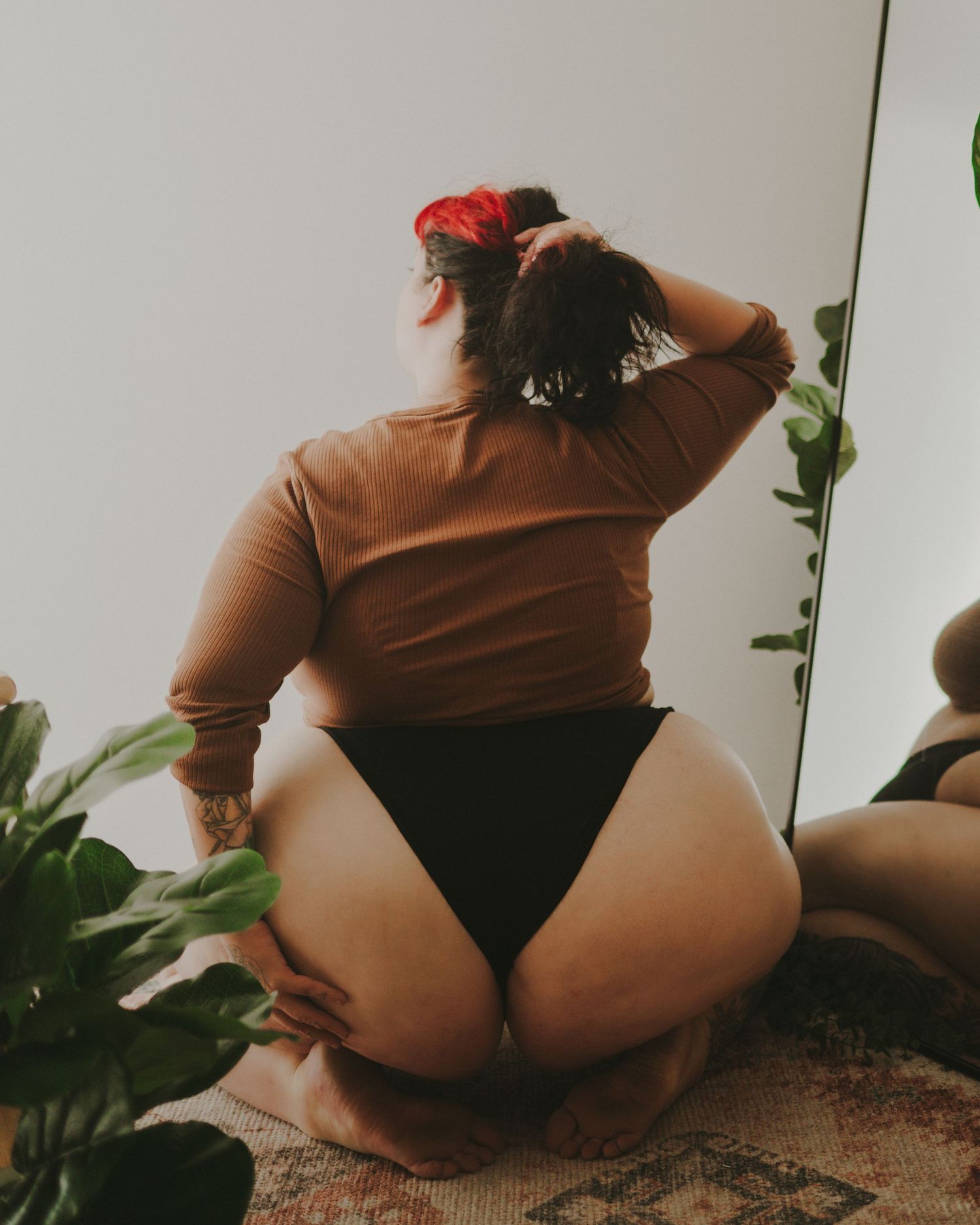 Back view of someone in a brown sweater and black bottoms crouching near indoor plants on a rug.