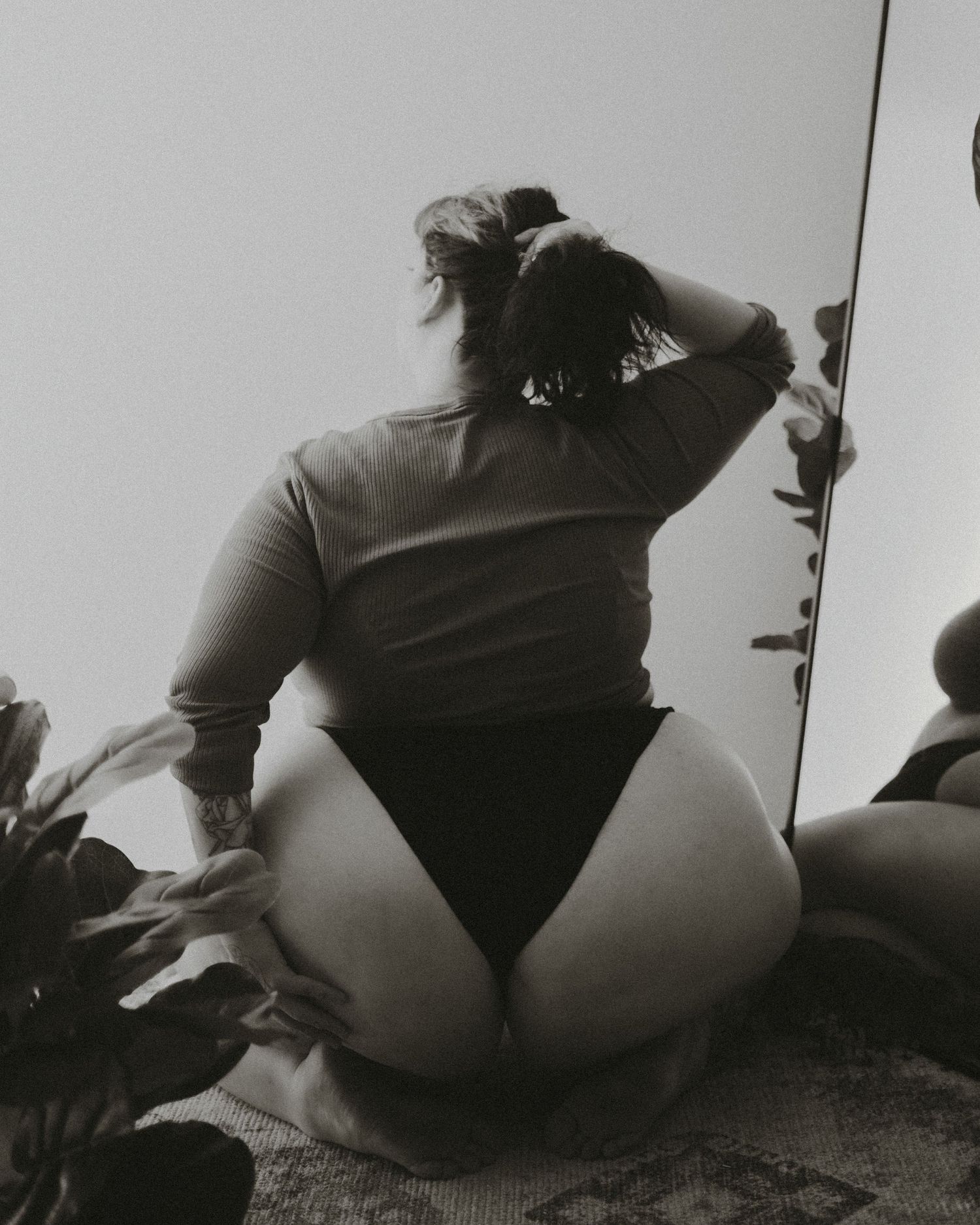 Black and white artistic photo showing person in a leotard crouching near potted plants against a white wall.