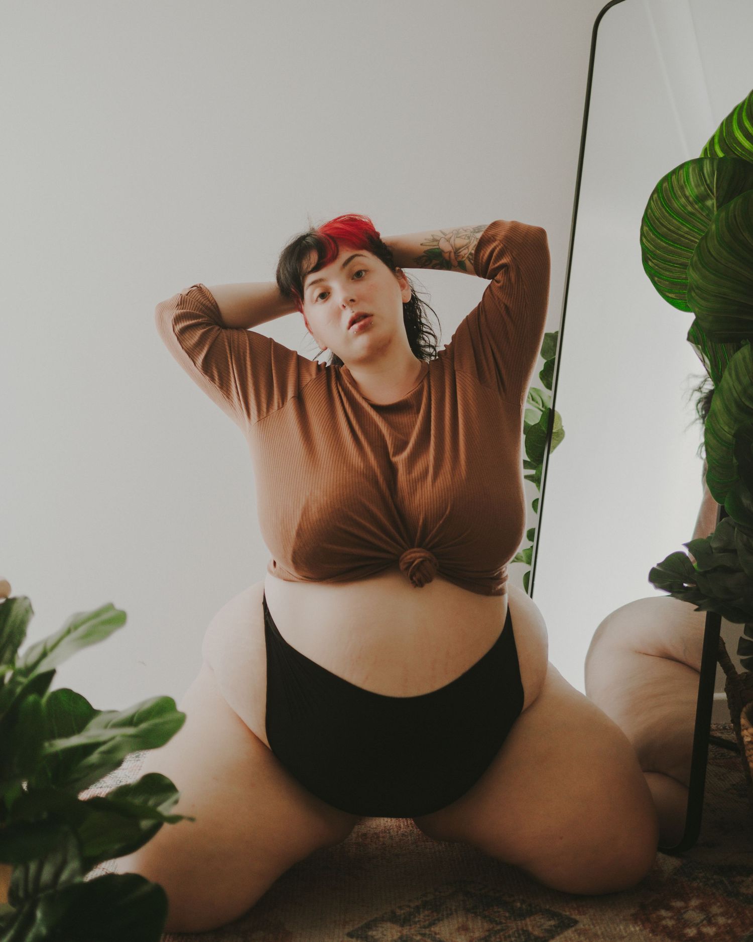 A model in brown knotted top and black shorts posing with houseplants against a white wall.