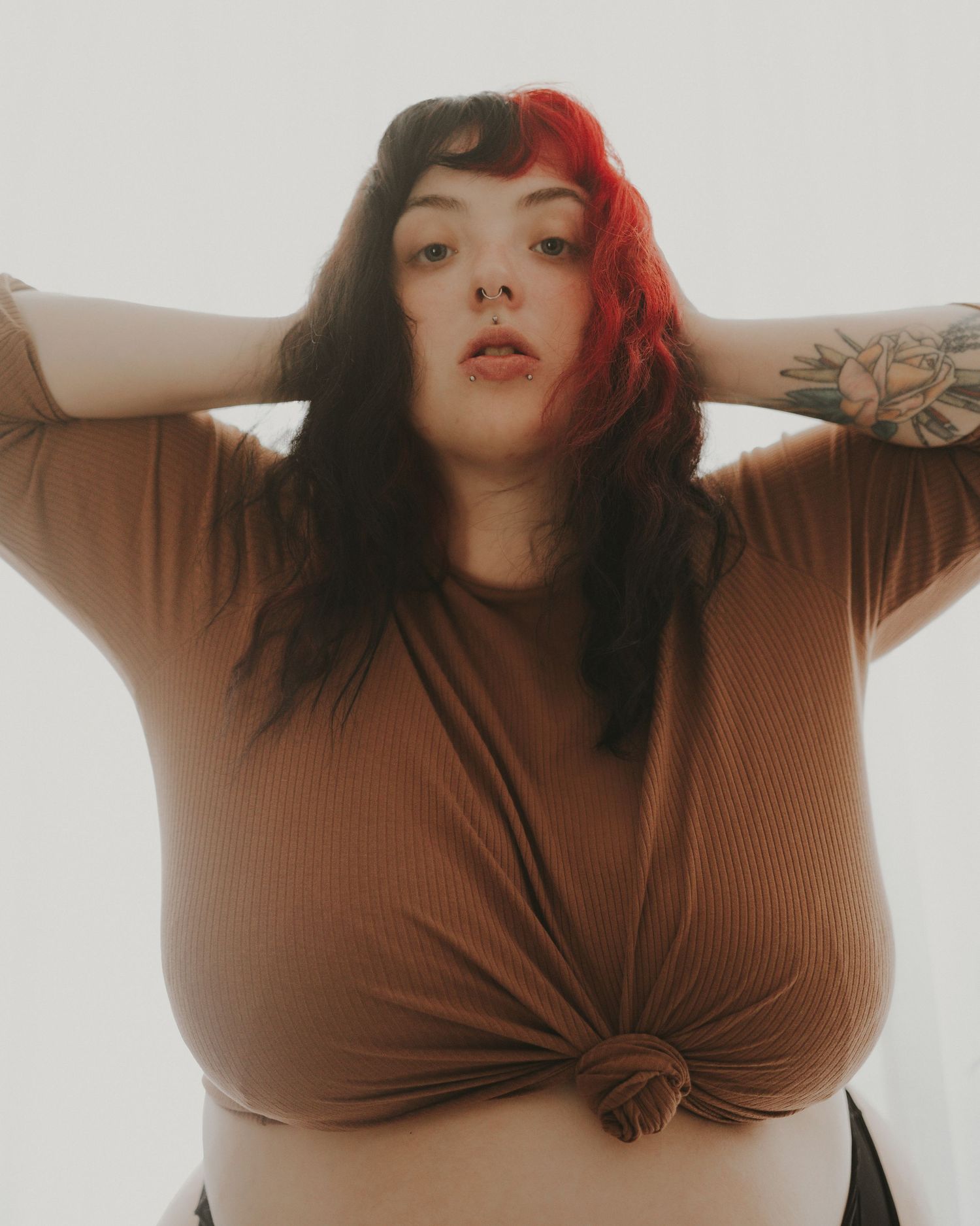 A portrait in warm lighting shows a figure in a brown knotted top with half-dyed black and red hair against a light backdrop.