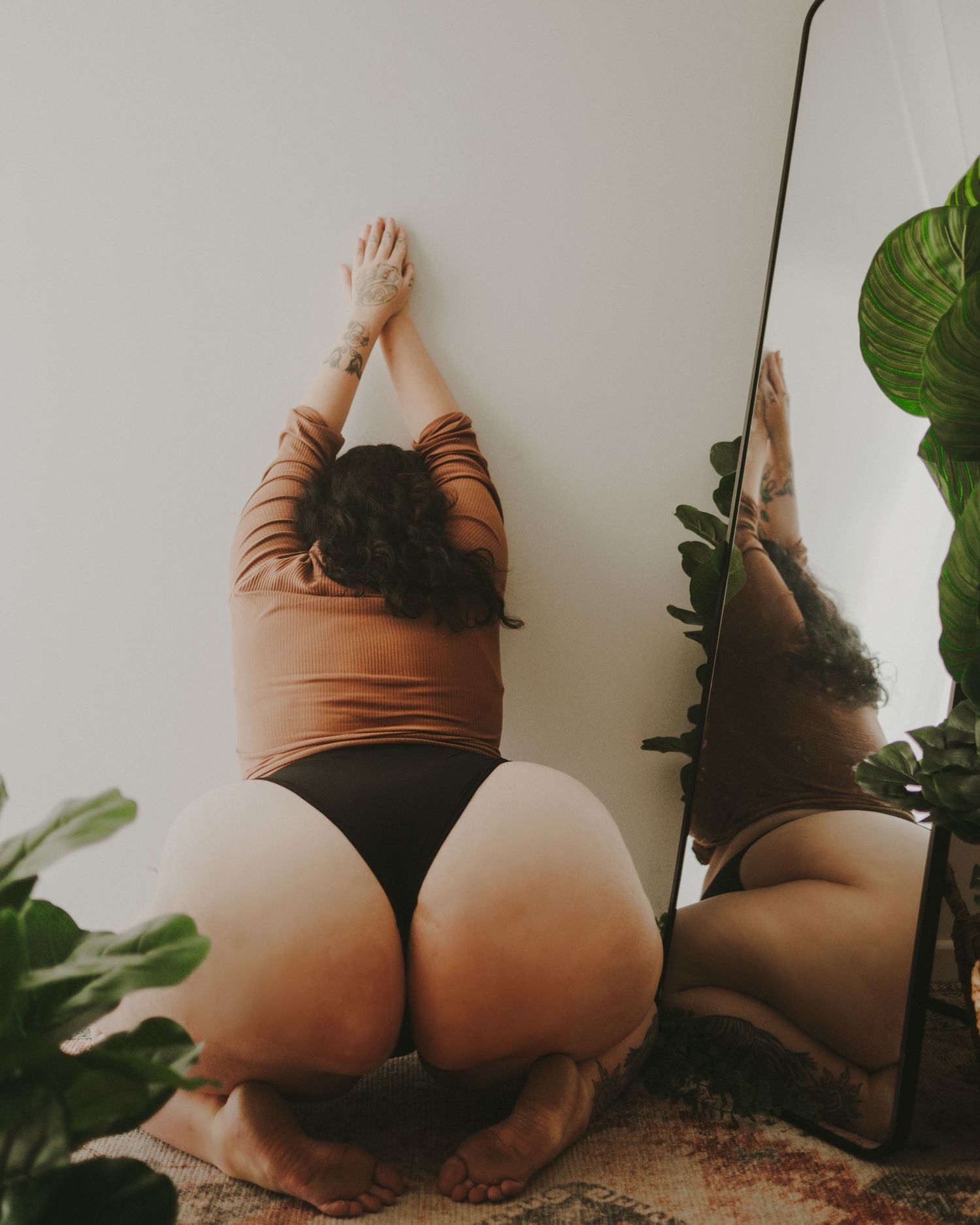 A person in brown top and dark bottoms stretches against a white wall with a mirror and potted plants nearby.