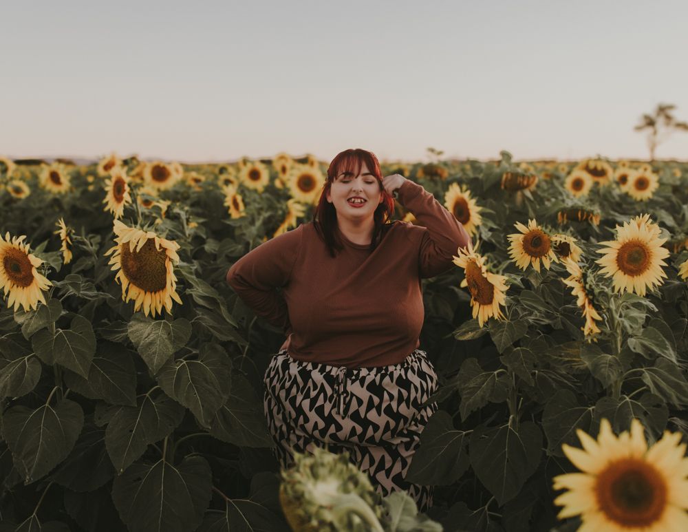 Person in brown sweater joyfully posing in a blooming sunflower field.