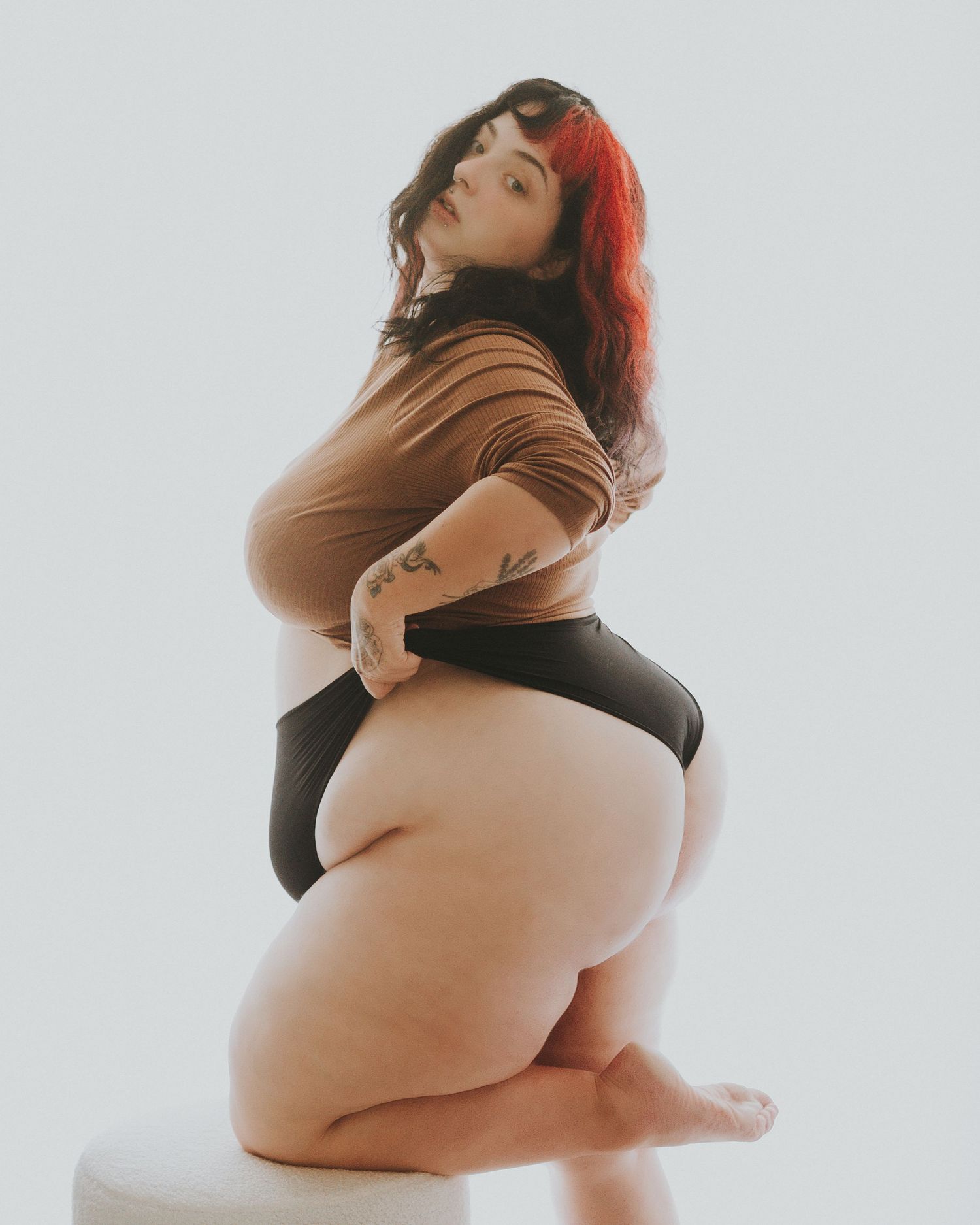 Boudoir style portrait showing a plus size model in brown bodysuit posing against white background.