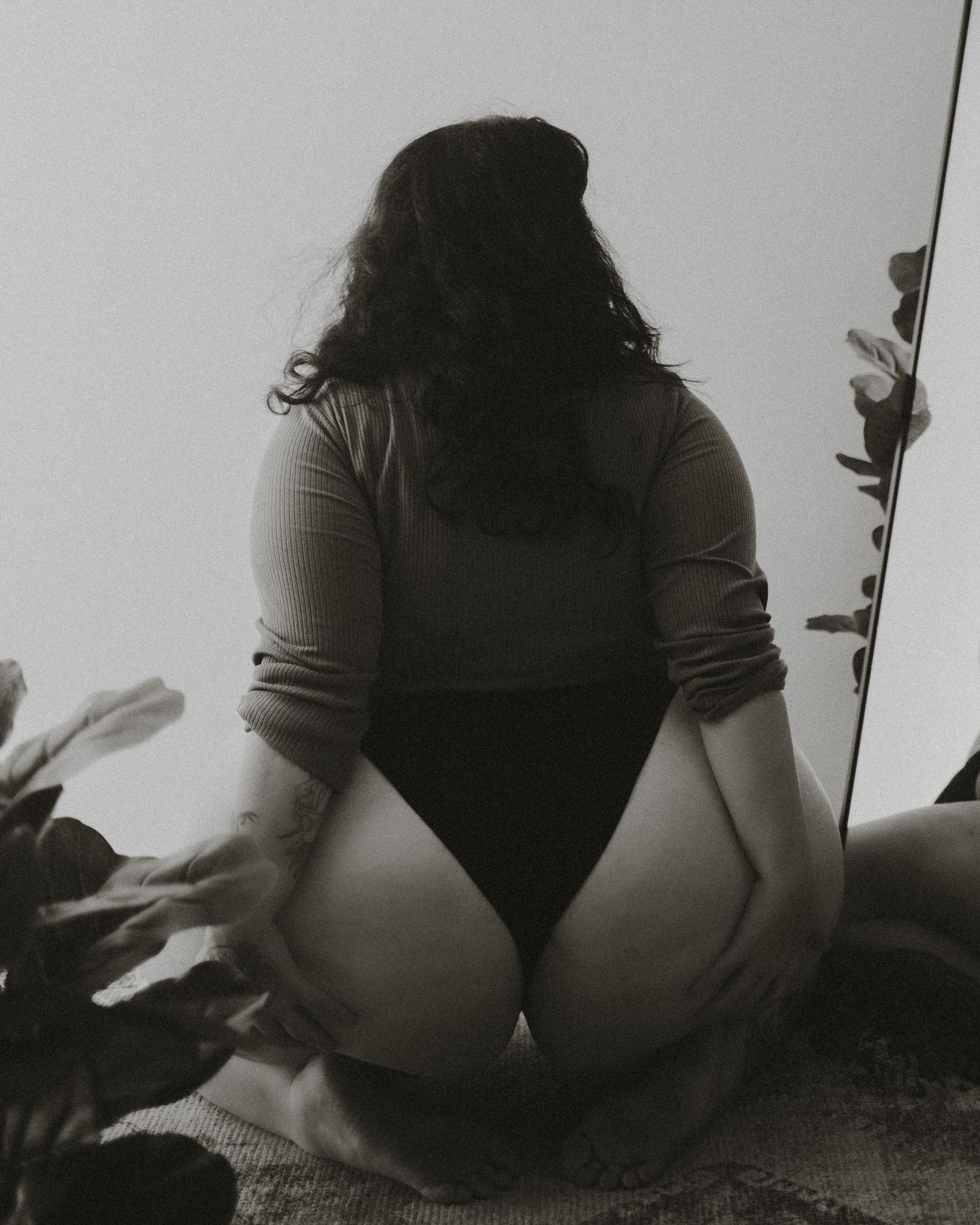 Black and white artistic boudoir photo showing a silhouette in intimate bodysuit sitting among shadows and plants.