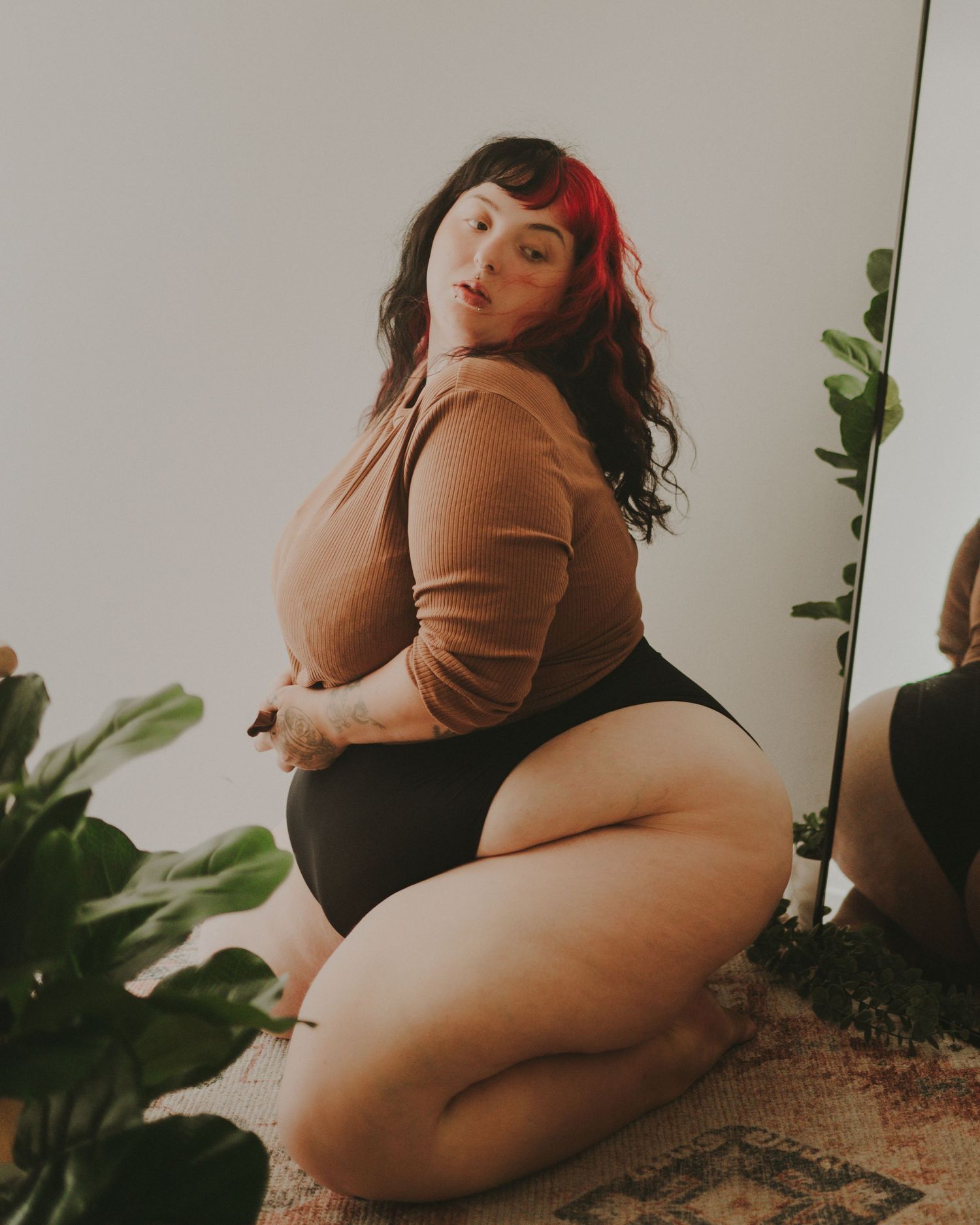 A plus size model in a tan sweater poses playfully near houseplants against a white wall.