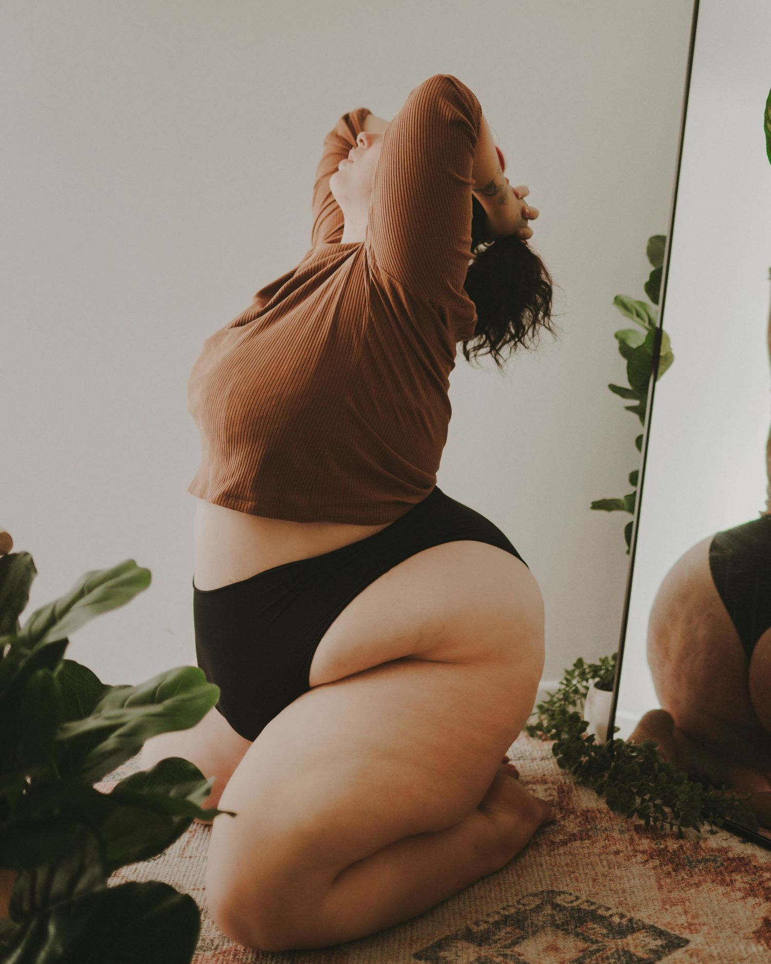 A plus size model in brown top and black underwear poses artistically with houseplants in natural lighting.