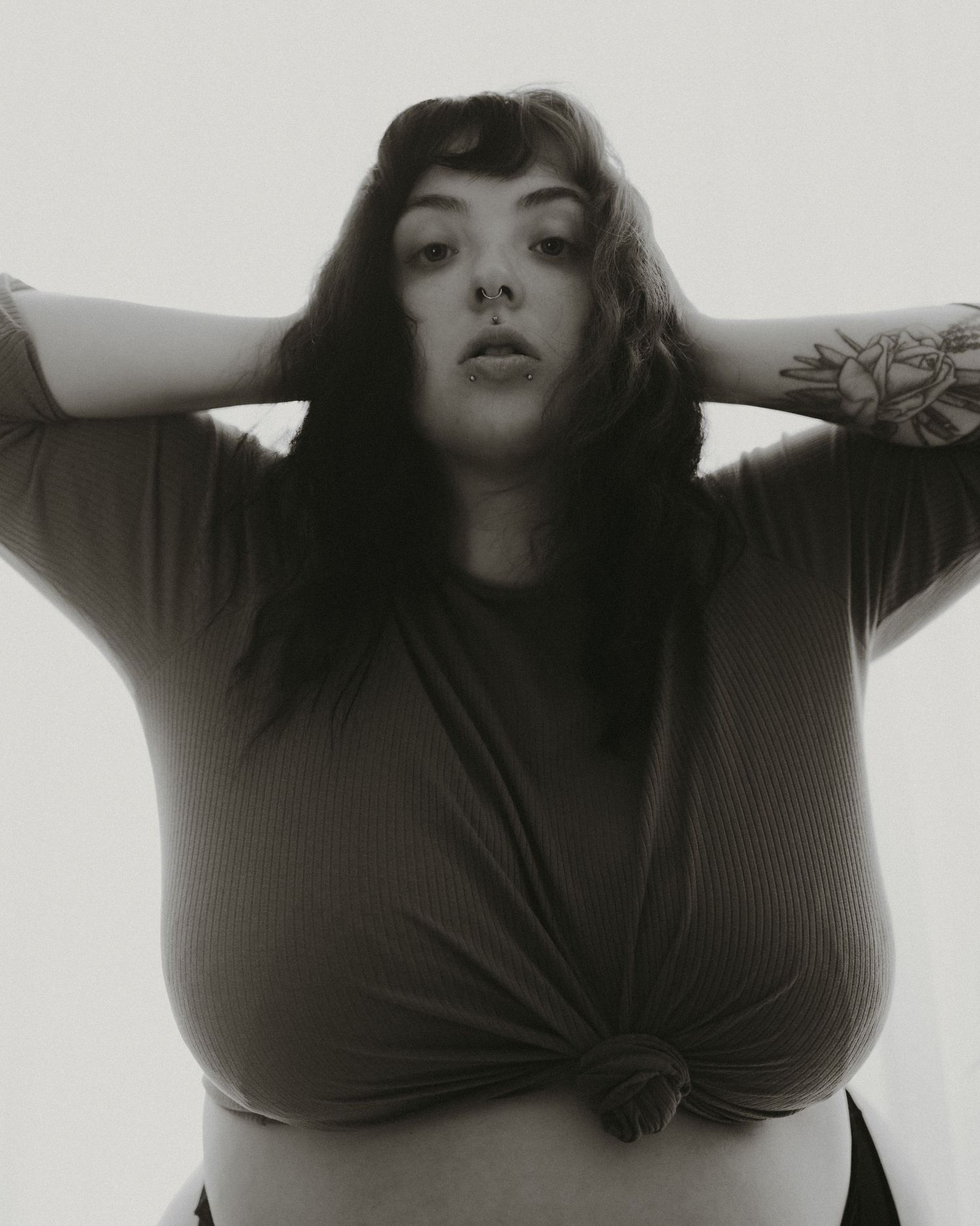 Black and white artistic portrait showing dark-haired figure in knotted shirt posing with hands raised behind head.
