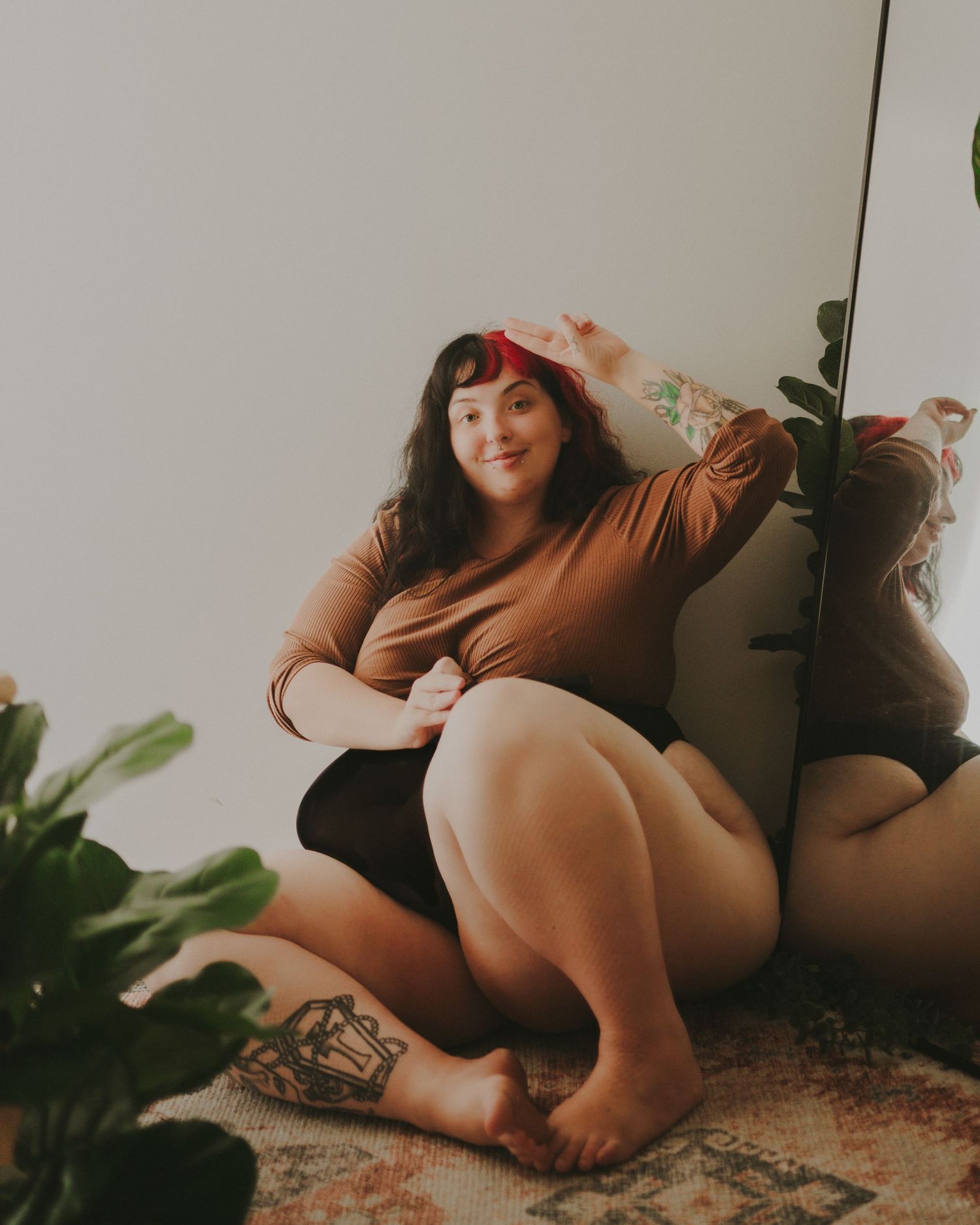 Plus size model in brown wrap top lounges casually against wall with potted plants in relaxed indoor photoshoot setting.