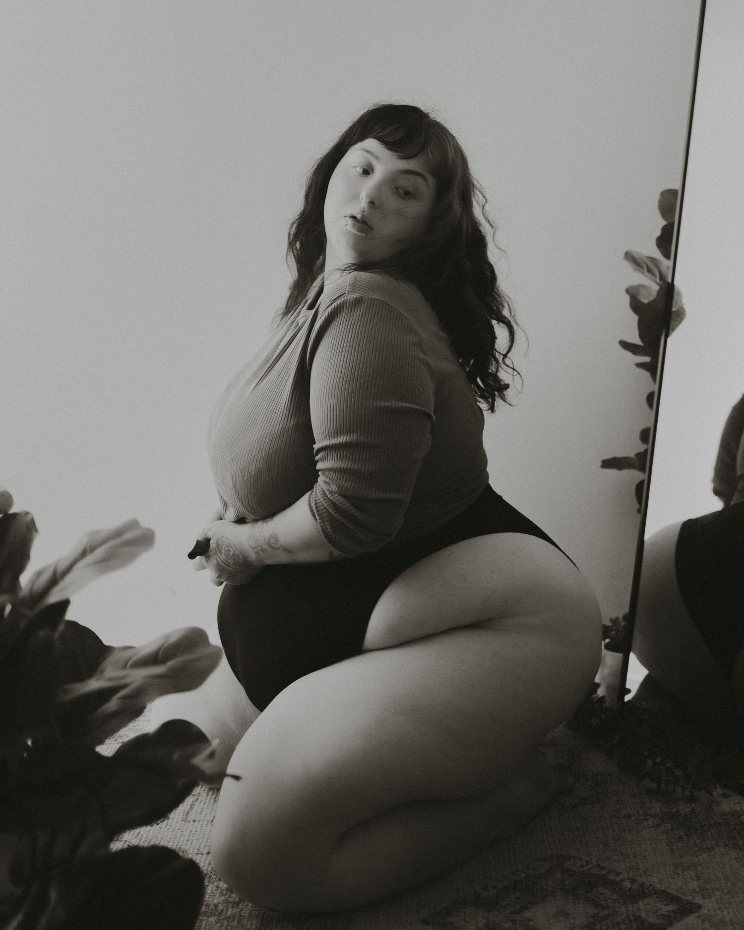 Monochromatic boudoir photo featuring a woman in knitwear posing artistically with indoor plants.