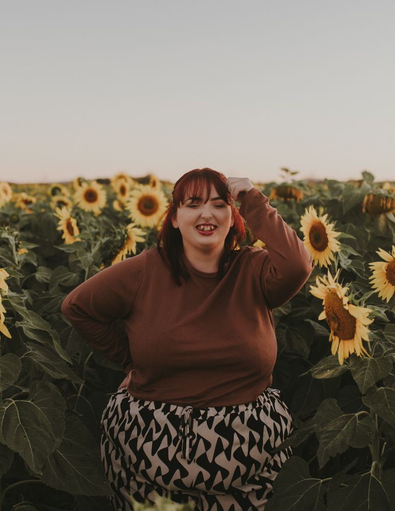 Person wearing brown sweater and patterned skirt standing among tall sunflowers.