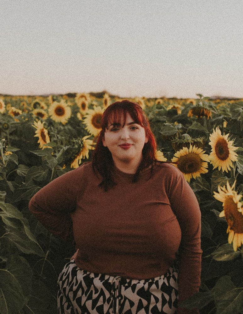 Person wearing brown sweater and geometric print skirt poses in sunflower field at sunset.