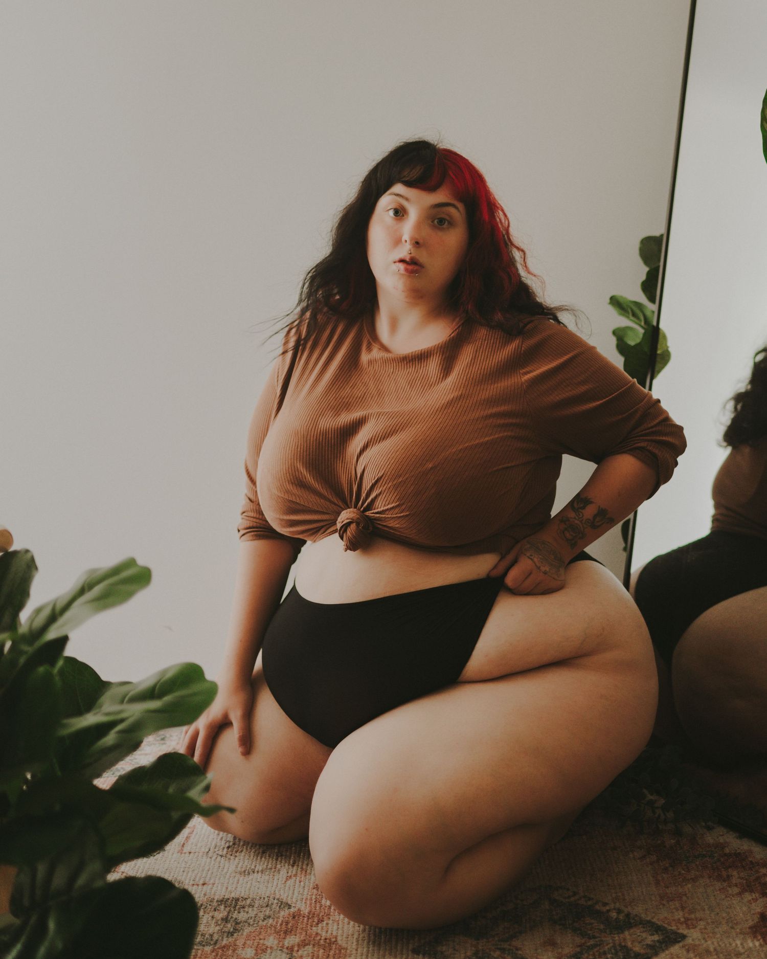 Figure in brown cropped top and black bottoms poses with plants in minimalist indoor setting with natural lighting.