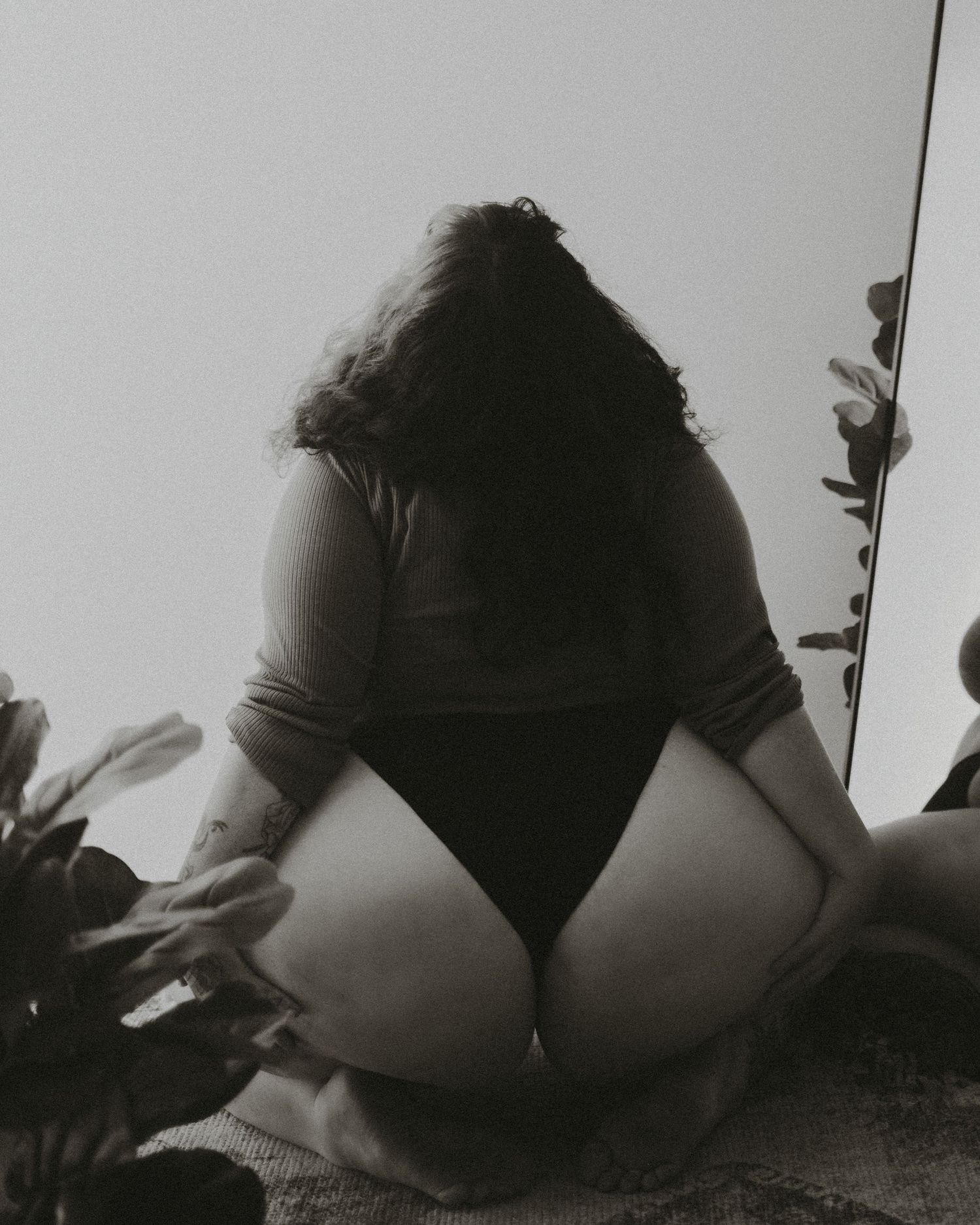 Black and white artistic boudoir photograph showing the back of someone in dark lingerie sitting with plants.