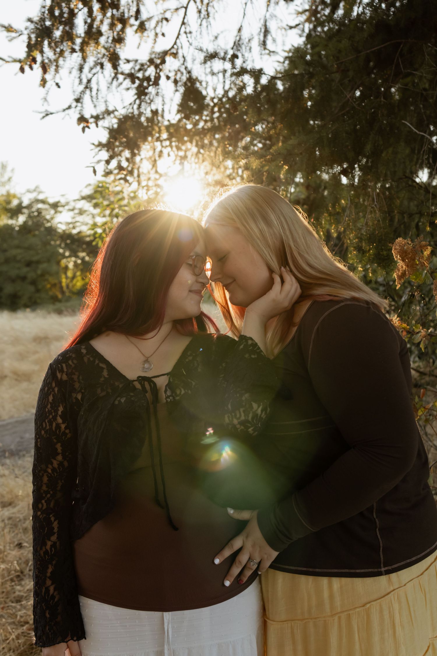 Two women share a tender forehead-to-forehead moment outdoors with golden sunlight streaming through trees behind them.