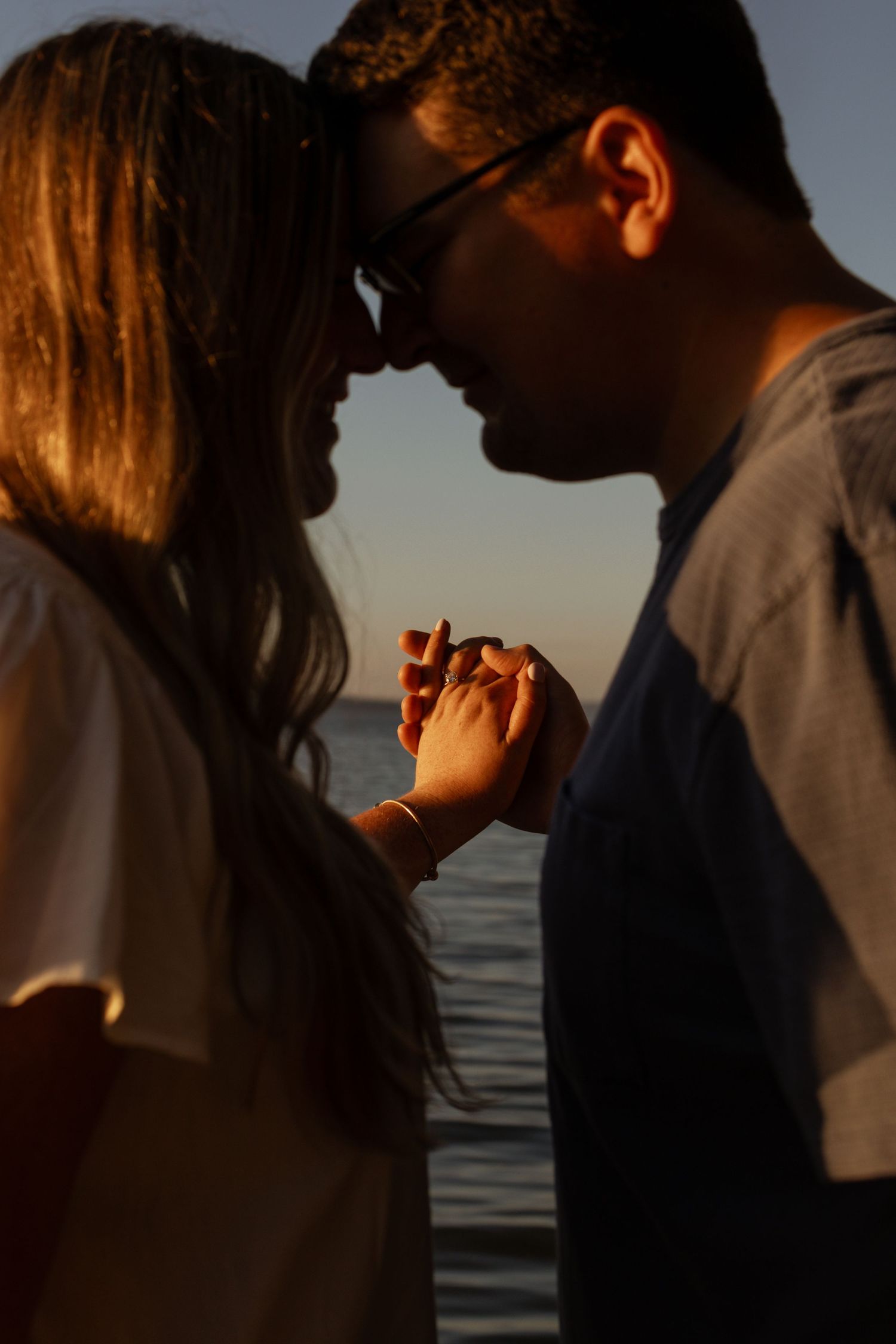 Romantic couple holding hands and touching foreheads at sunset by the ocean.