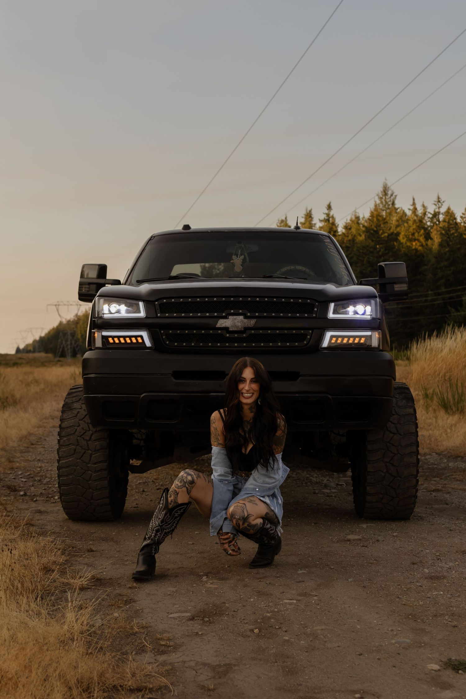 Woman posing in front of a lifted black Chevrolet truck on a dirt road at dusk.