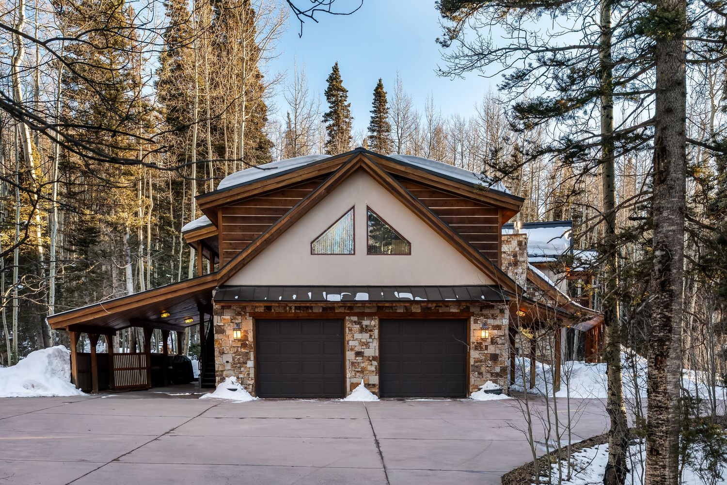 Snow-covered mountain cabin with stone accents, wooden siding, and two-car garage in a forest setting.