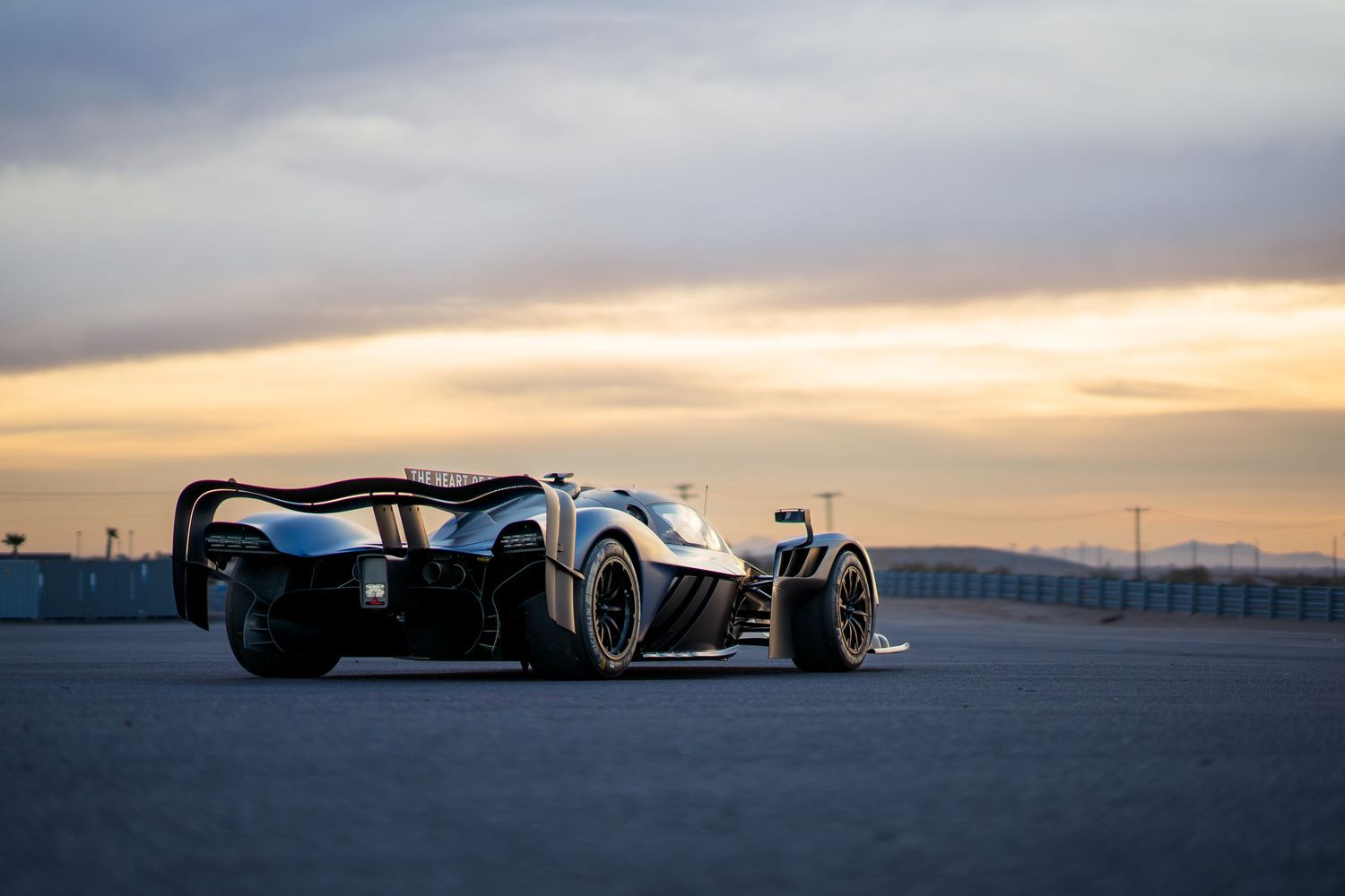 Blue racing prototype car silhouetted against dramatic sunset sky at a racetrack.
